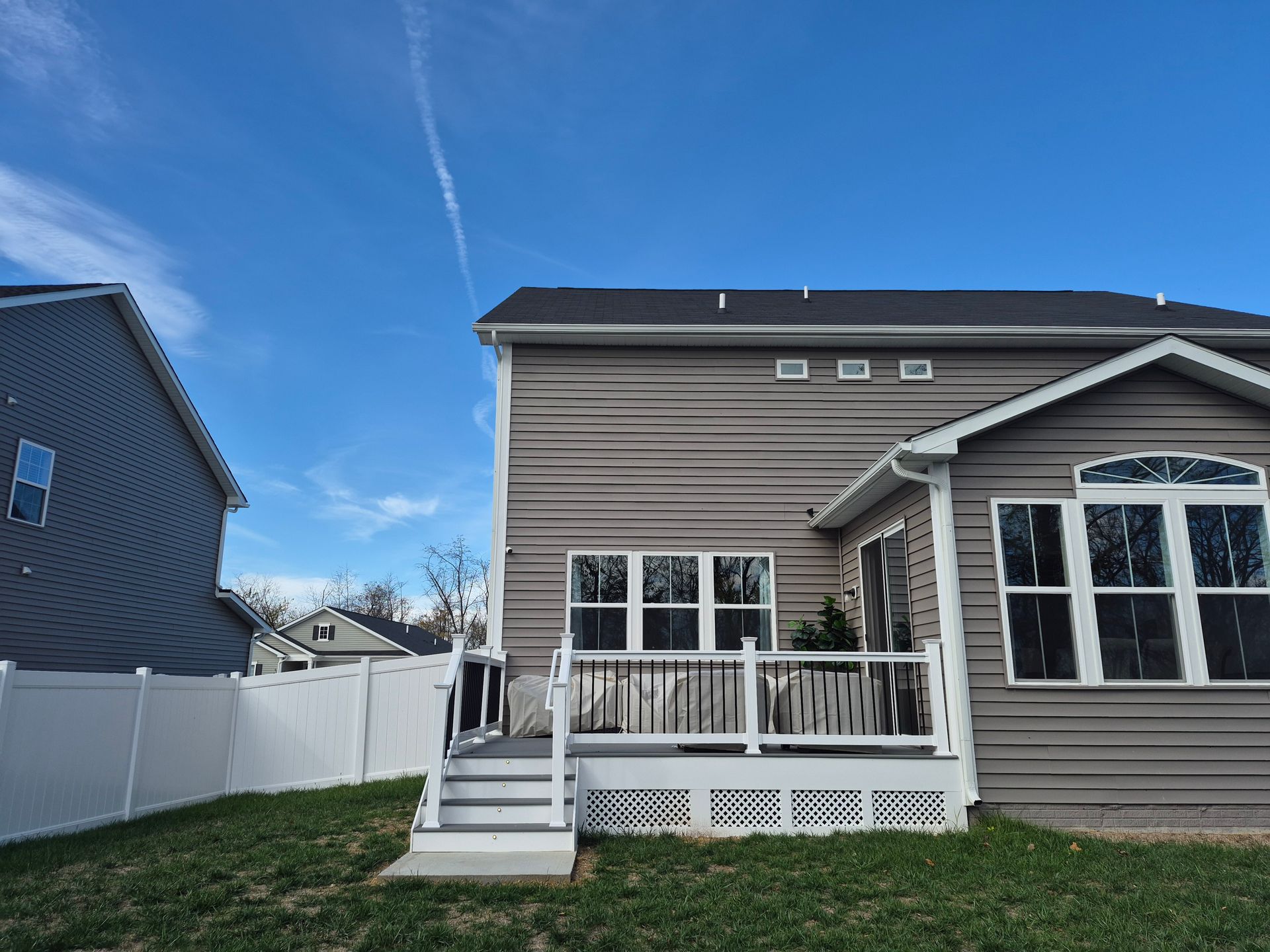 Backyard view of two-story houses, one with gray siding and a deck, the other with blue siding, against a blue sky.