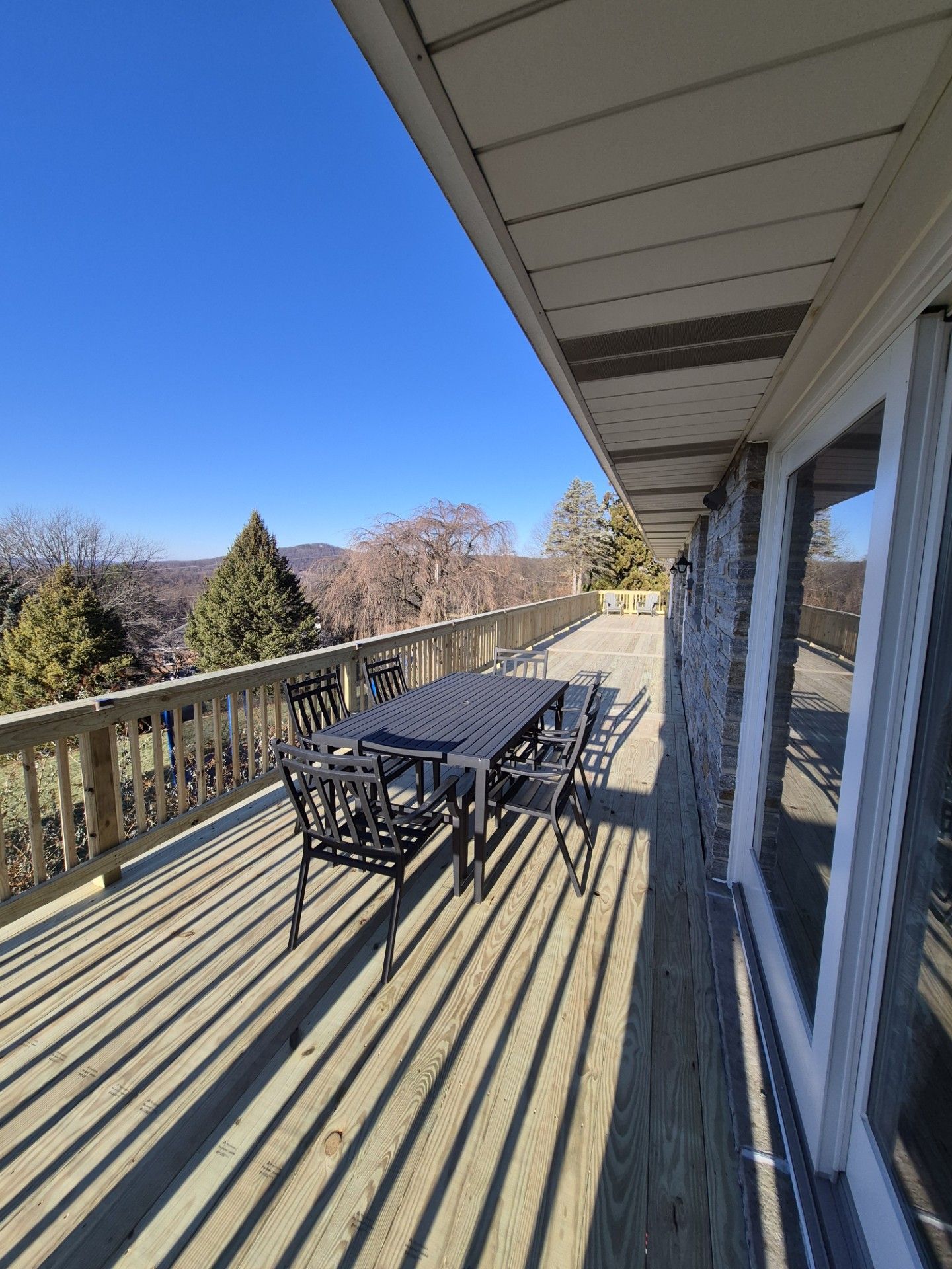 Wooden deck with a dark table and chairs, overlooking a landscape with trees under a blue sky.