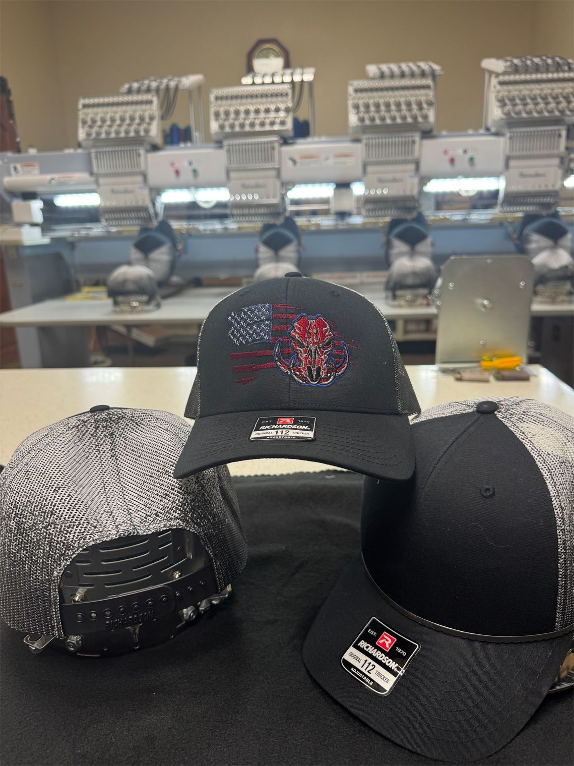 Three hats are sitting on a table in front of embroidery machines