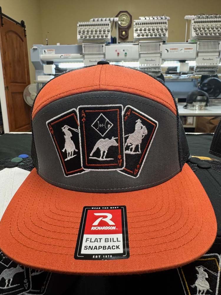 Orange and gray flat-bill snapback hat with playing card designs and a black mesh back, sitting in front of embroidery machines.
