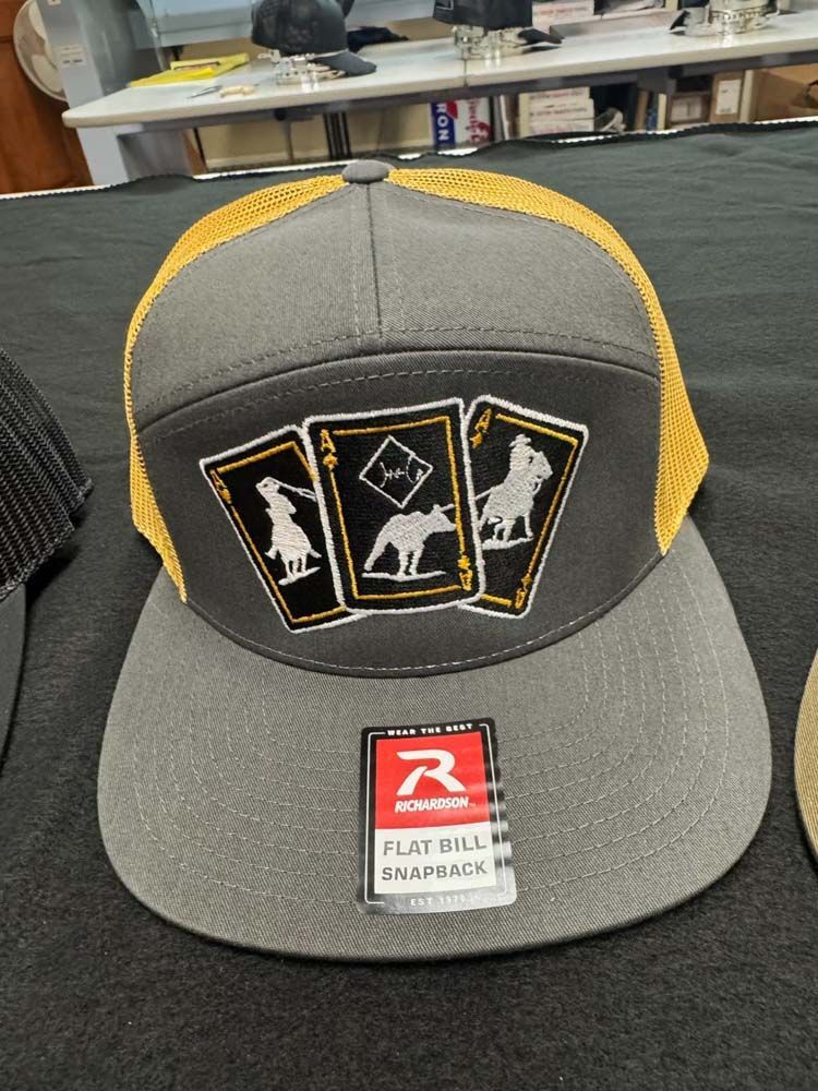 Grey and gold trucker hat with playing card design and a flat brim.