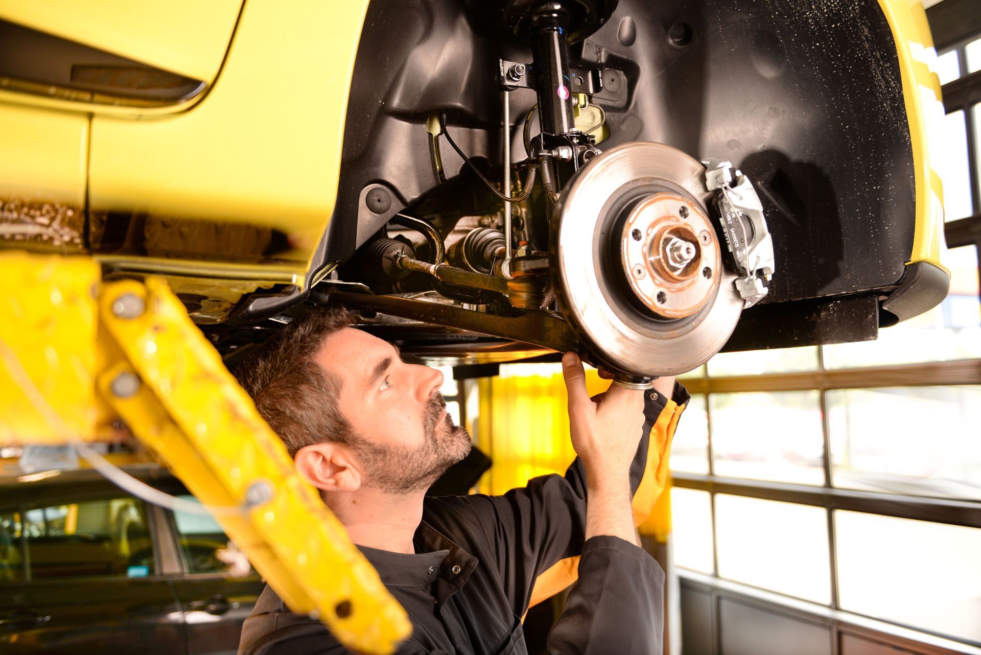 Mechanic inspecting car brakes, car raised on yellow lift.