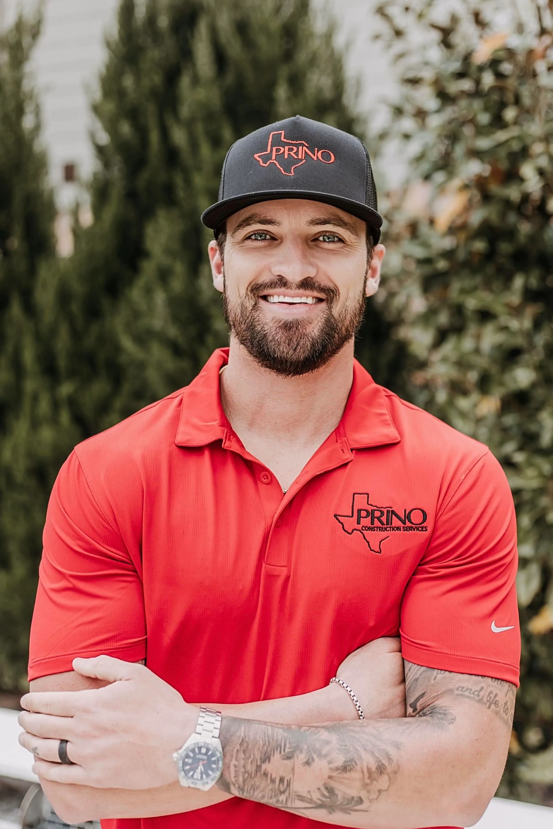 Man in a red polo and black cap standing outdoors with arms crossed, smiling at the camera.