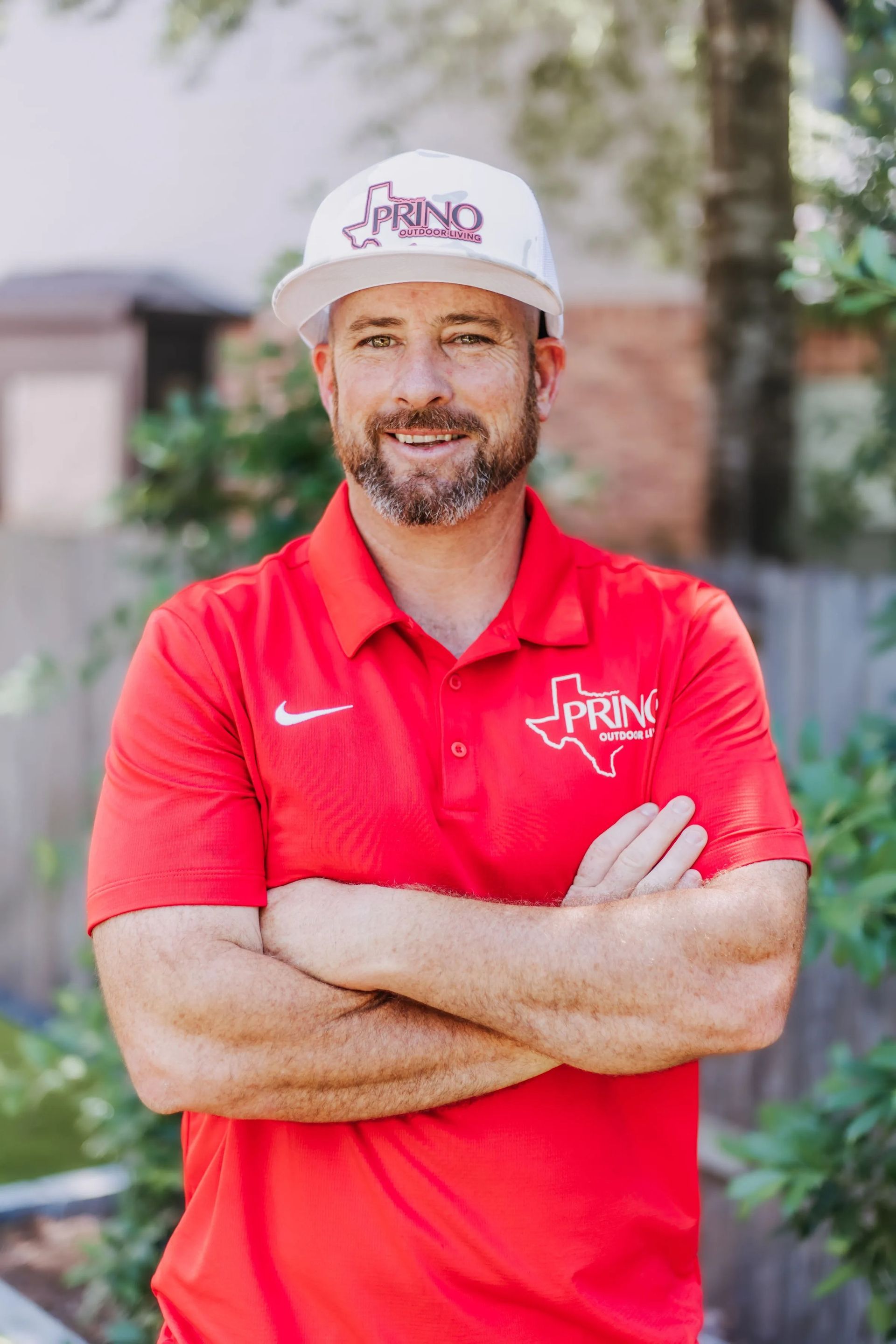 Man in red polo and white cap stands outdoors with arms crossed, smiling.