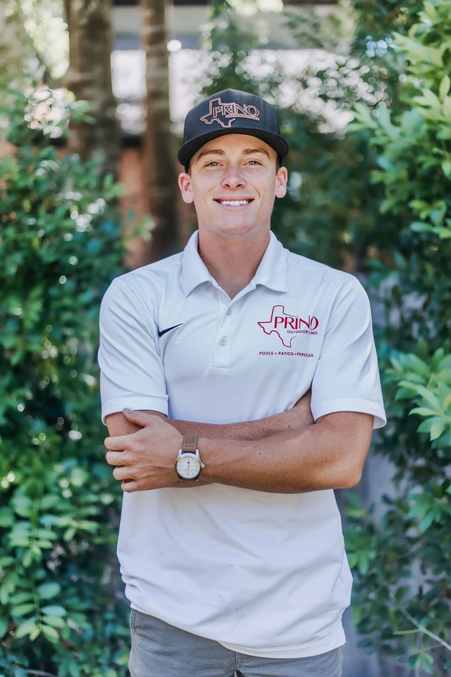 Smiling person in a black cap and white polo, standing outdoors with arms crossed in front of greenery.