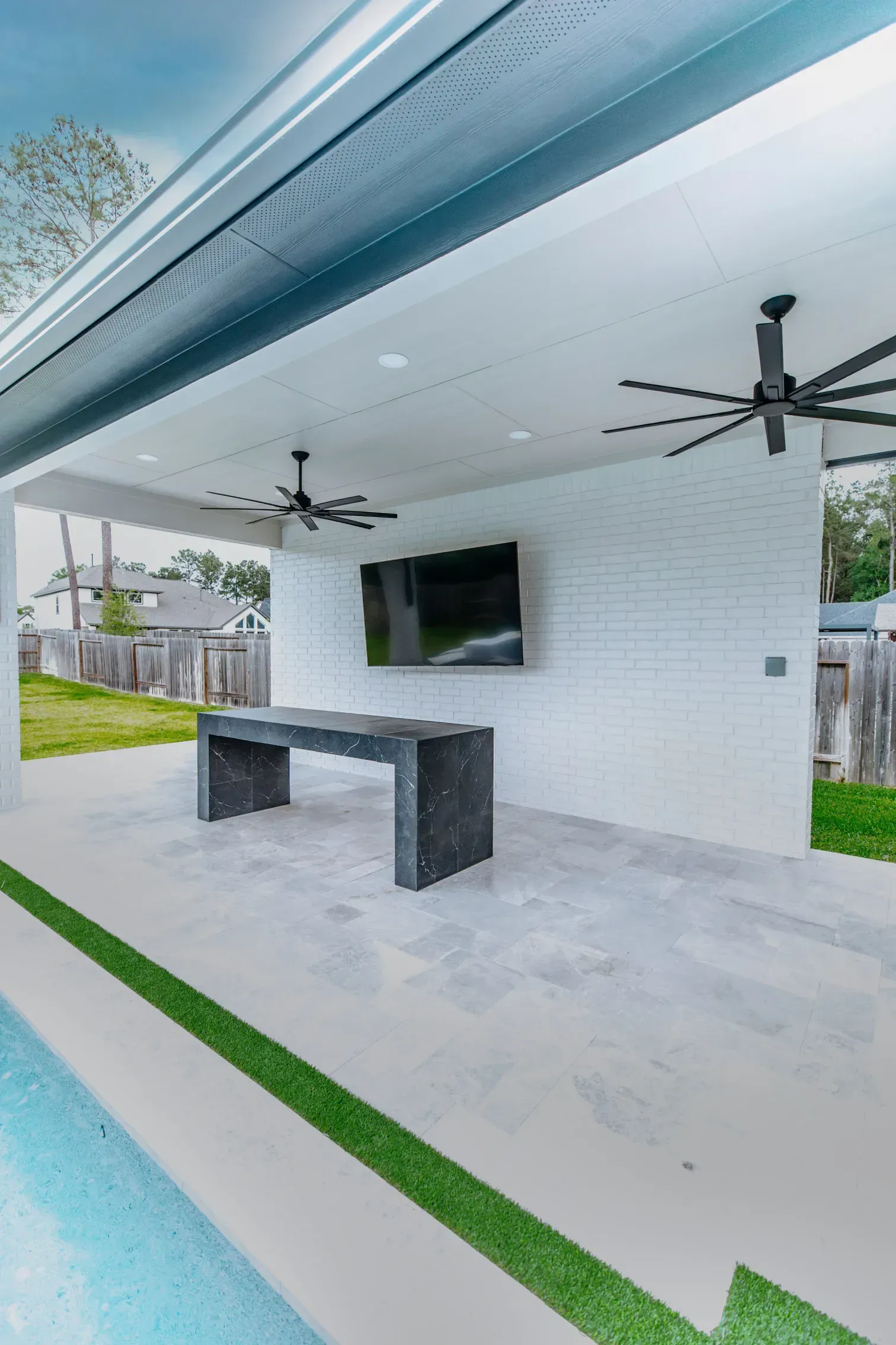 Covered patio with ceiling fans, wall-mounted TV, and a stone table beside a pool.