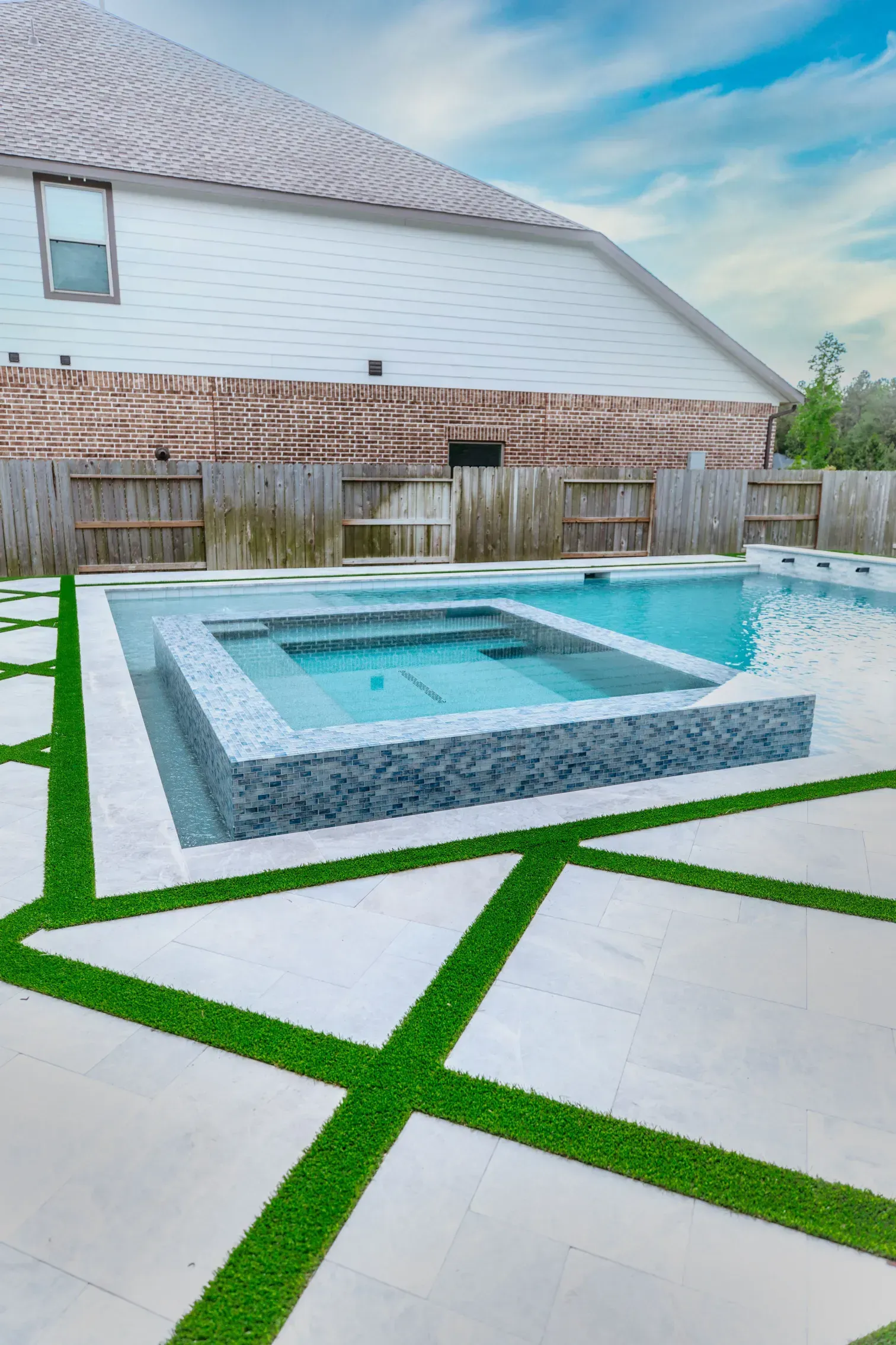 Backyard pool with white pavers and green grass inlays beside a white house