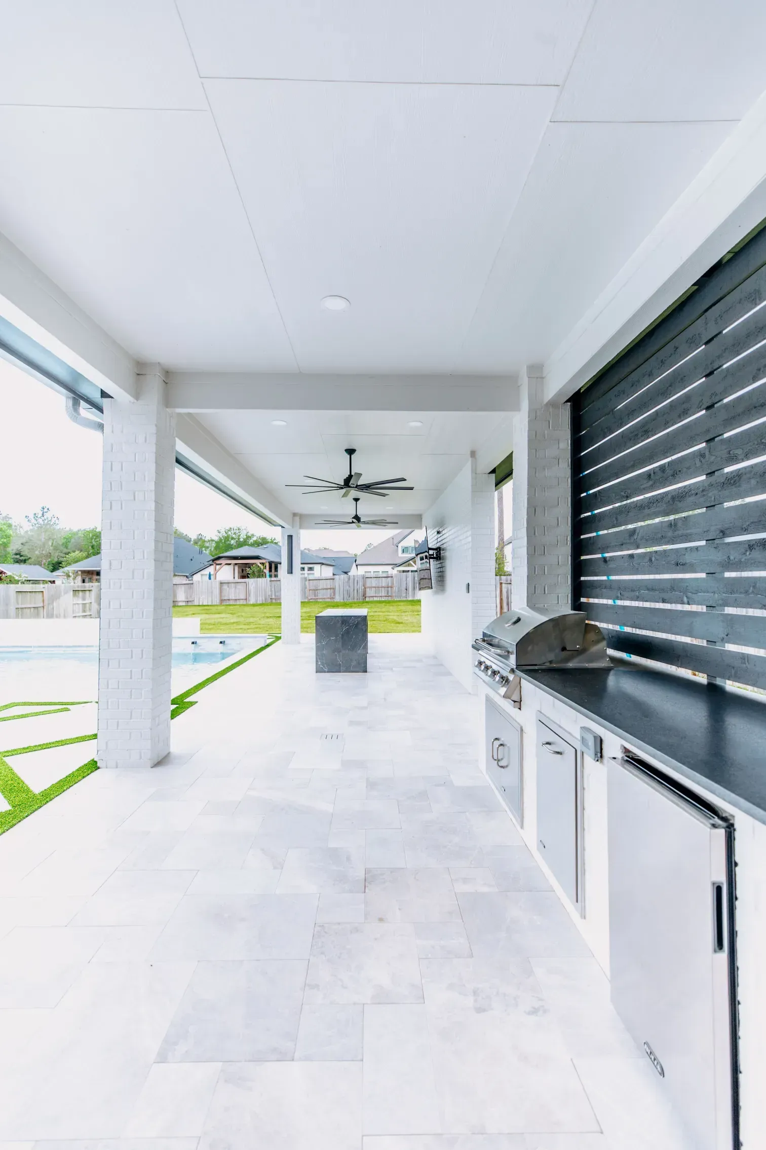 Covered patio with white tile floor, ceiling fan, grill, and black louvered privacy wall overlooking a yard