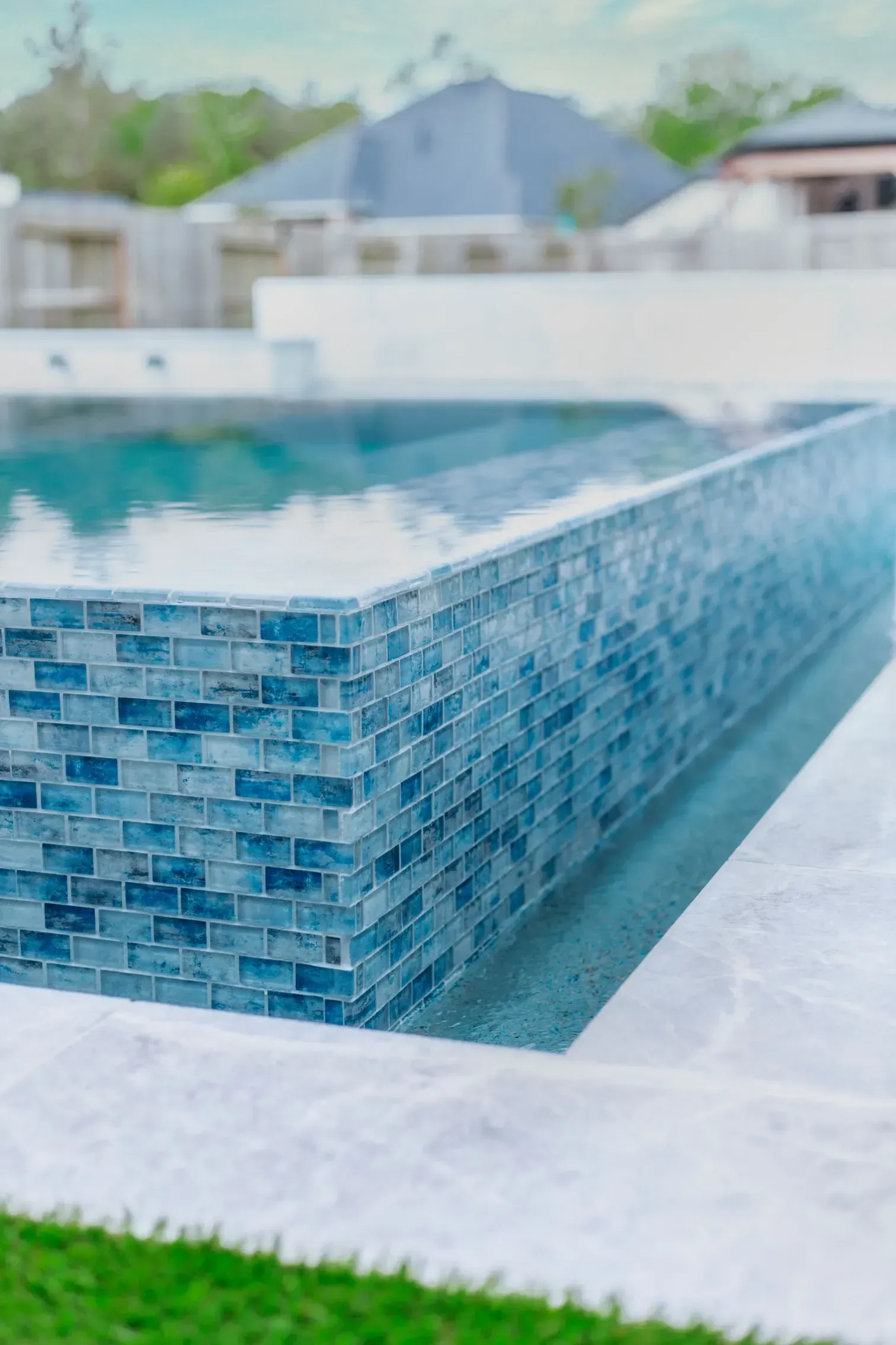 Blue-tiled pool corner with white coping and a blurred house in the background