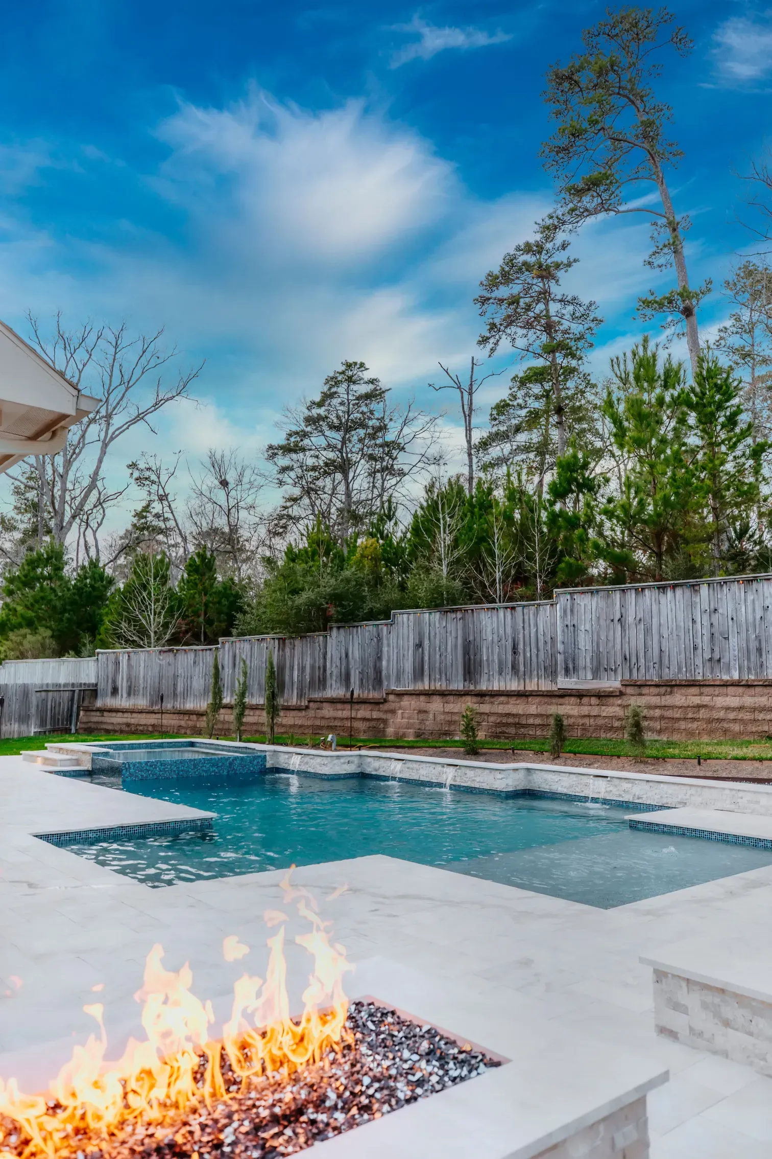 Backyard pool with a lit fire pit, white deck, and tall wooden fence under a blue sky