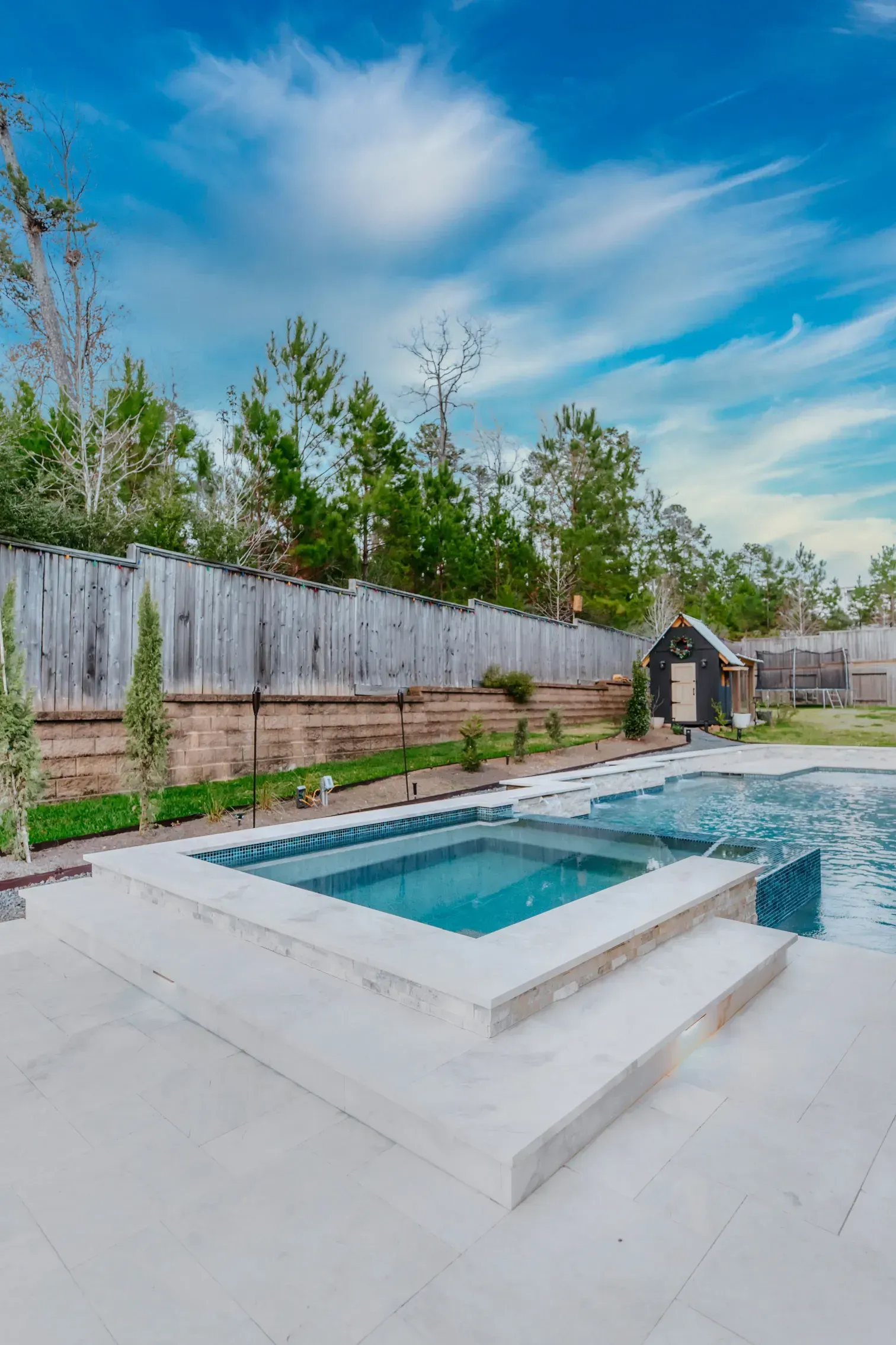 Backyard pool with raised spa, white patio, wooden fence, and blue sky above