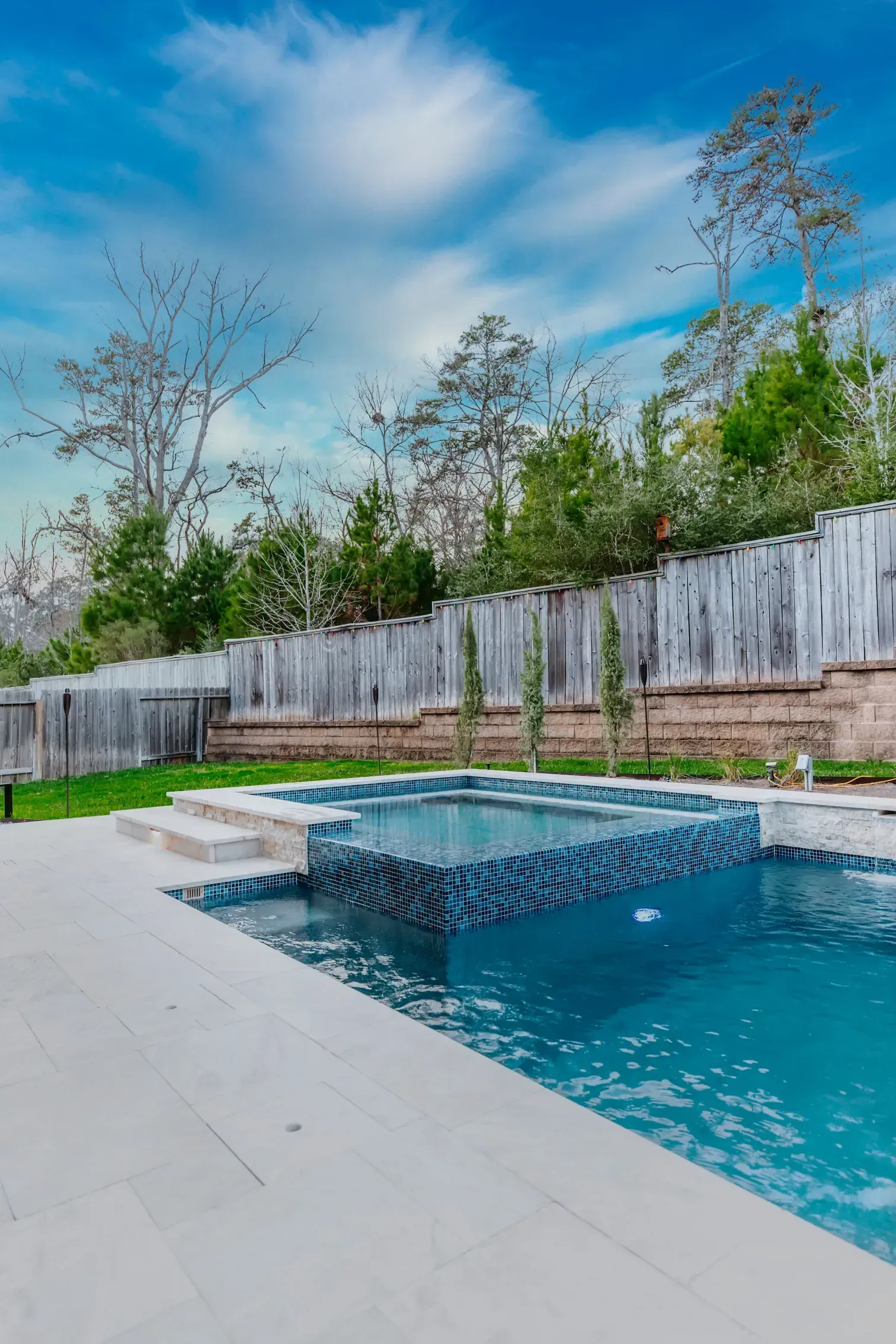 Backyard pool with a dark square water feature, white deck, wooden fence, and trees under a blue sky