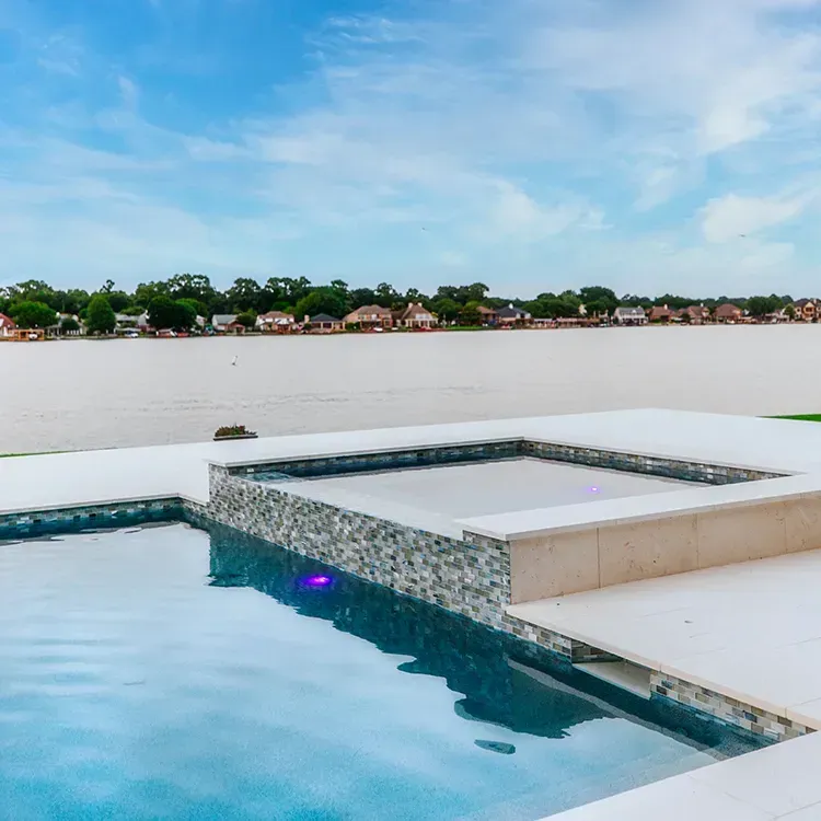 Infinity pool beside a river, with a tiled hot tub and distant shoreline under a blue sky