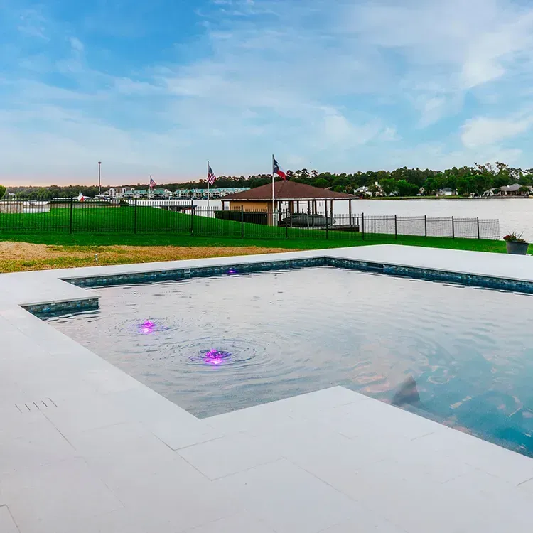 Modern backyard pool with a white deck, glowing purple lights, and a grassy waterfront view beyond