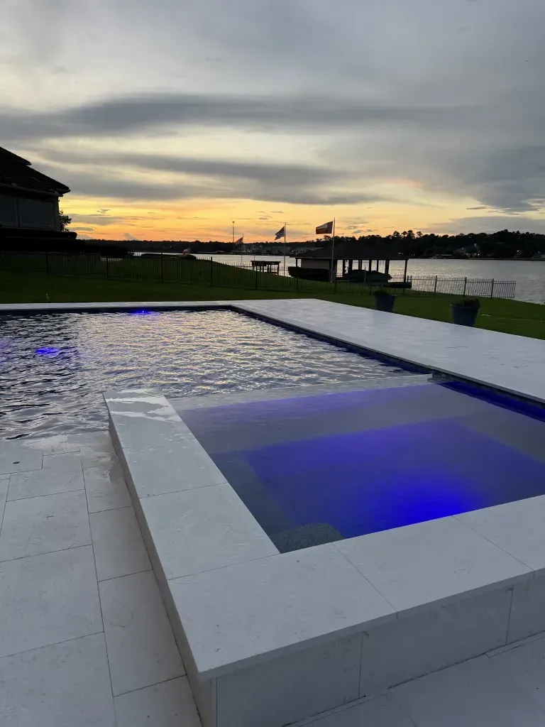 Modern pool at sunset with blue-lit spa, waterfront view, and cloudy sky