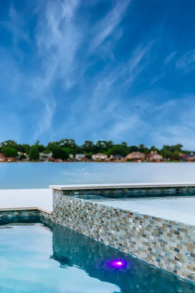 Infinity pool with mosaic edge reflecting a blue sky over a calm lake and distant houses