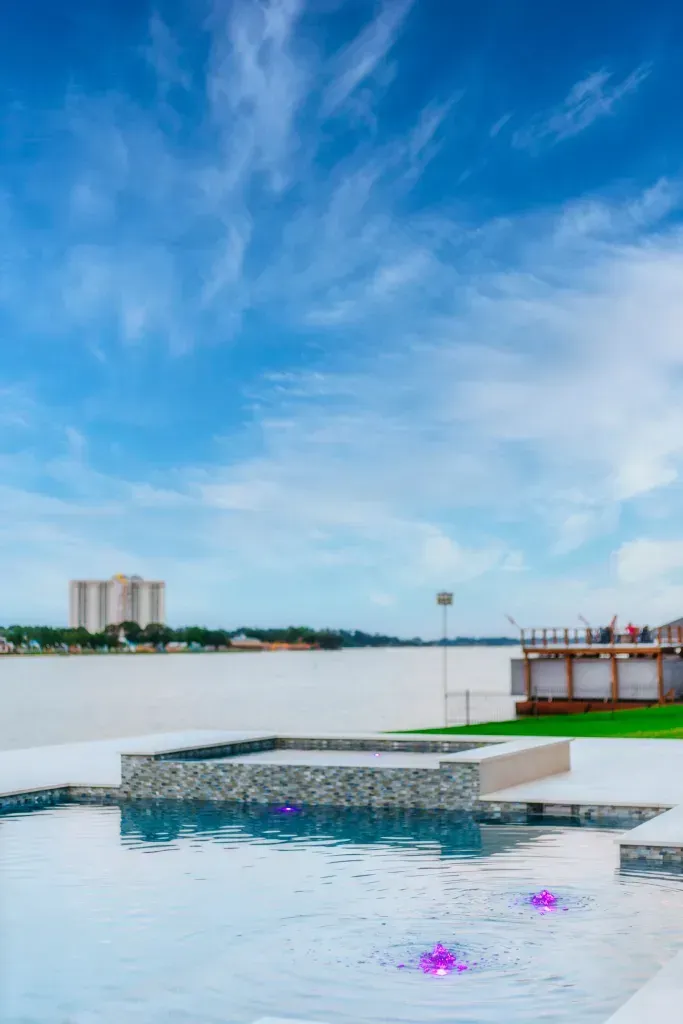 Infinity pool overlooking a calm waterfront under a blue sky, with a dock and distant buildings.