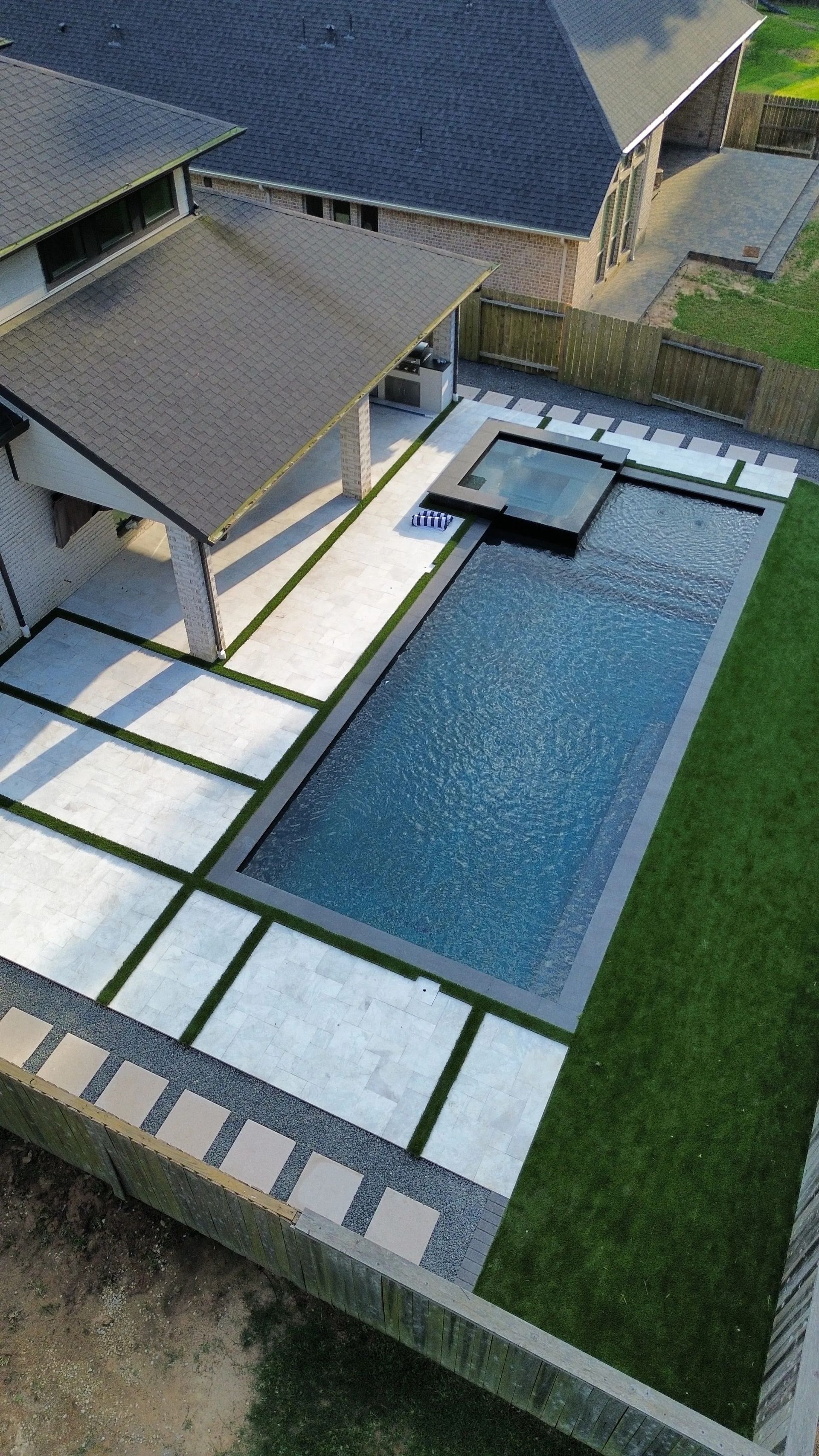 Aerial view of a modern backyard with a rectangular swimming pool, stone patio, and grass lawn.
