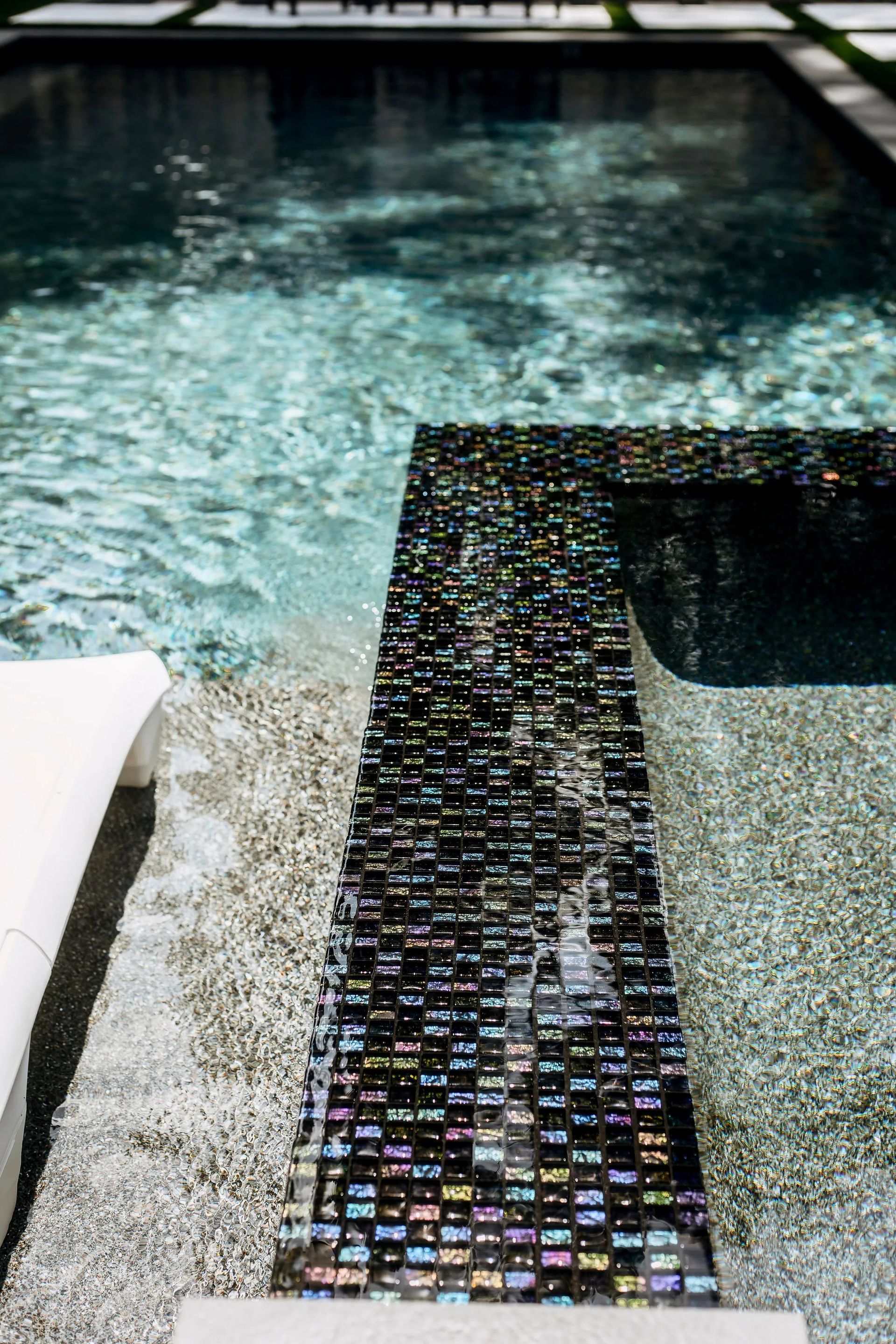 Mosaic-tiled pool ledge beside clear blue water with a white lounge chair nearby