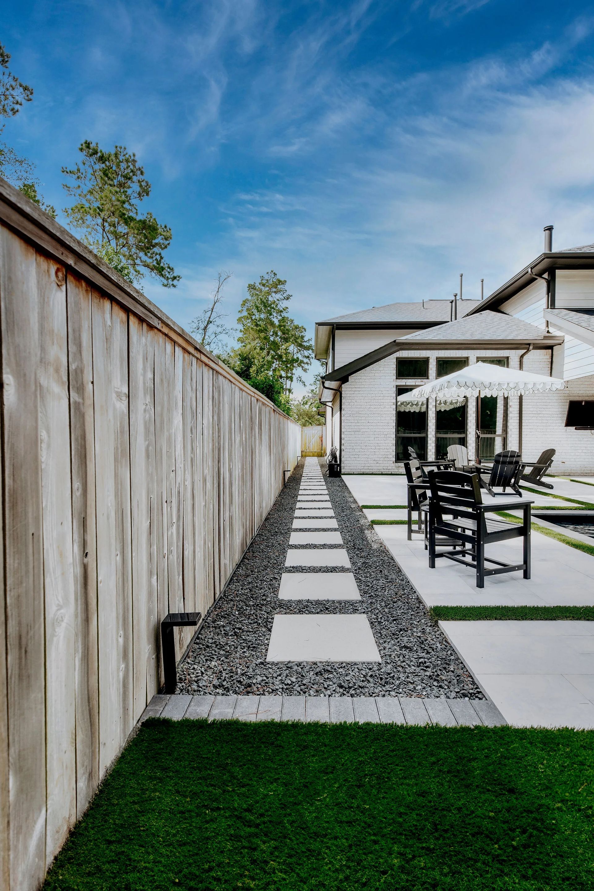 Narrow gravel path beside wooden fence leading to a patio with white house and outdoor chairs