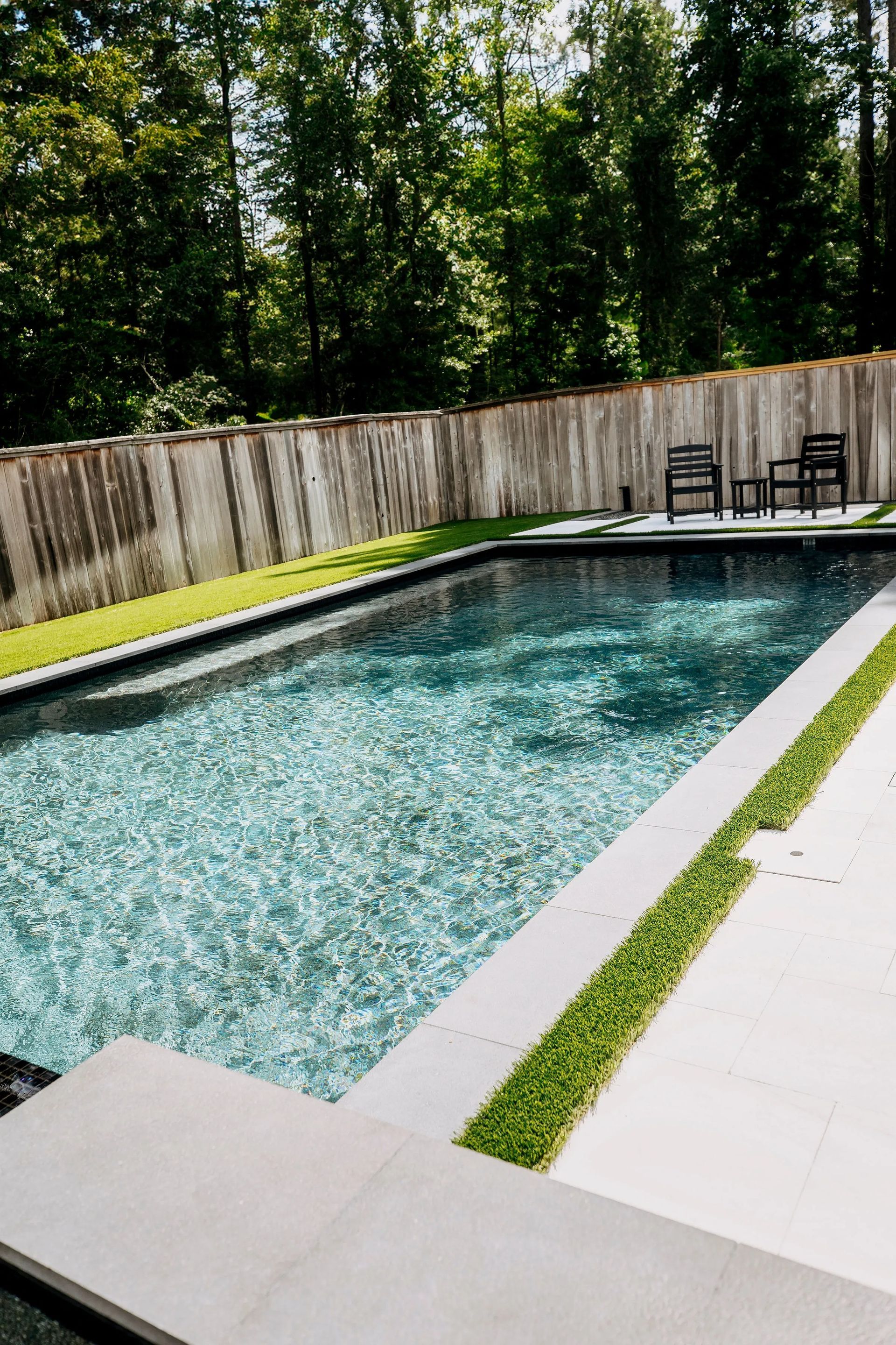 Rectangular backyard pool with blue water, white deck, wood fence, and lounge chairs by trees