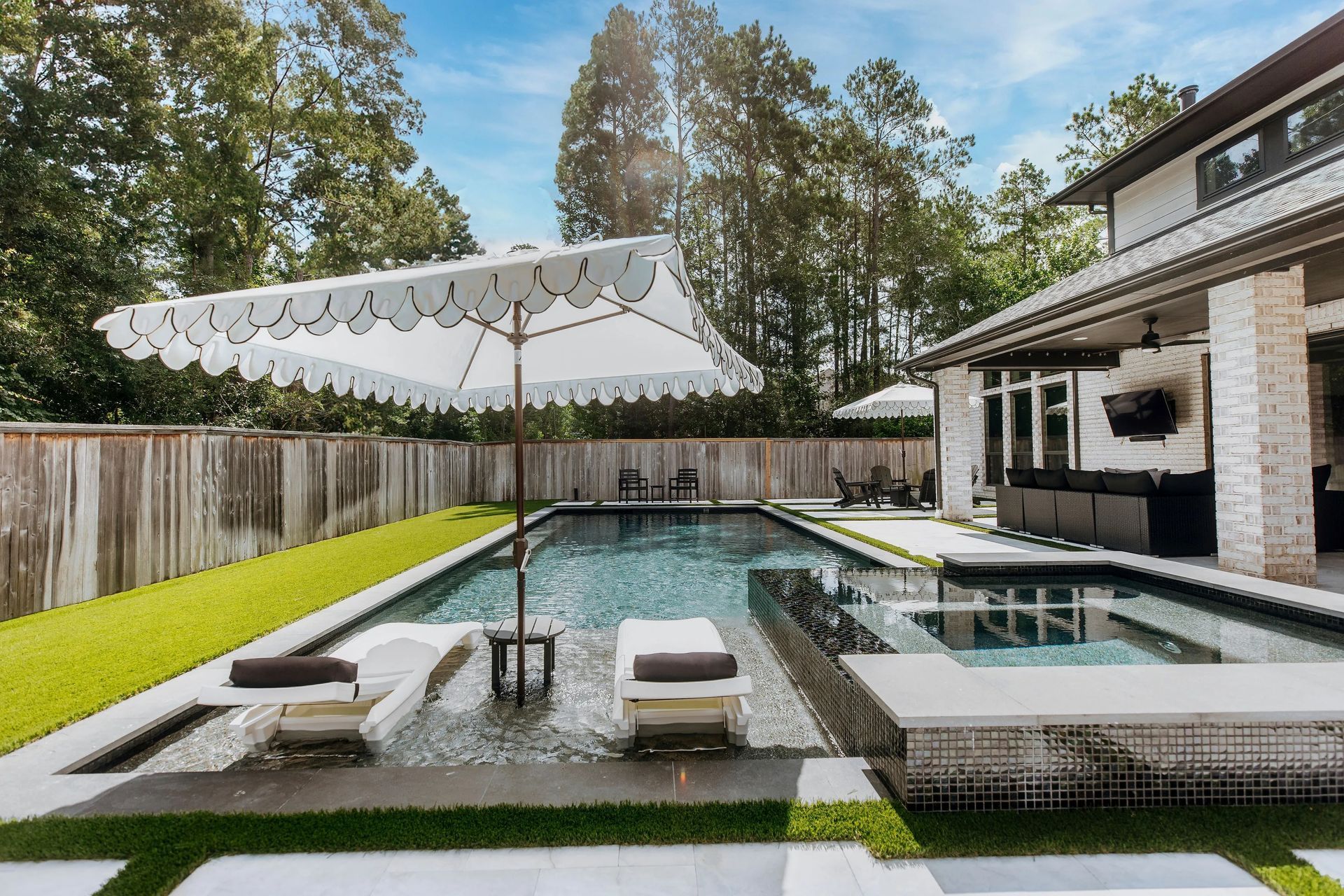 Modern backyard pool with lounge chairs, white umbrella, and a two-story house beside a wooden fence.