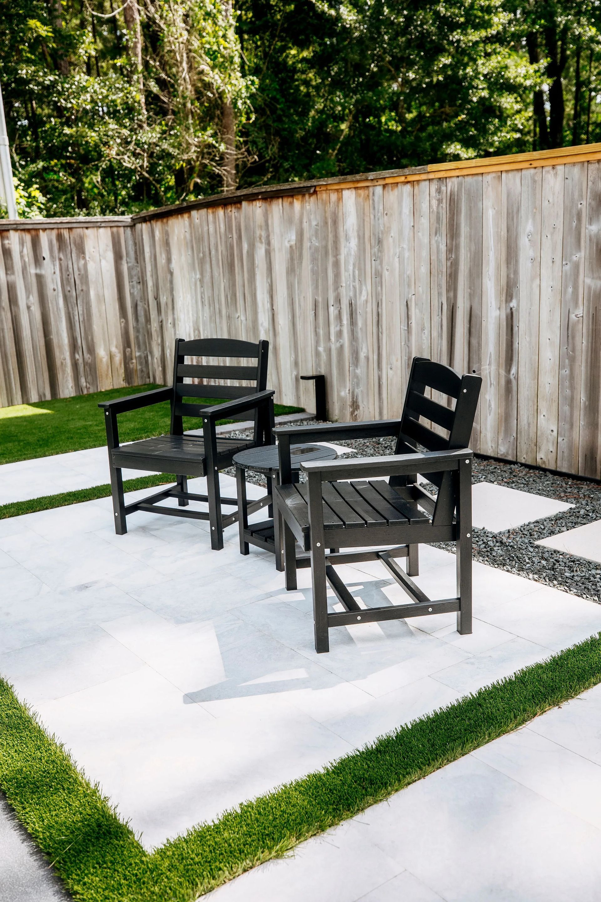Black patio chairs around a small table on a white concrete patio beside a wooden fence and trees