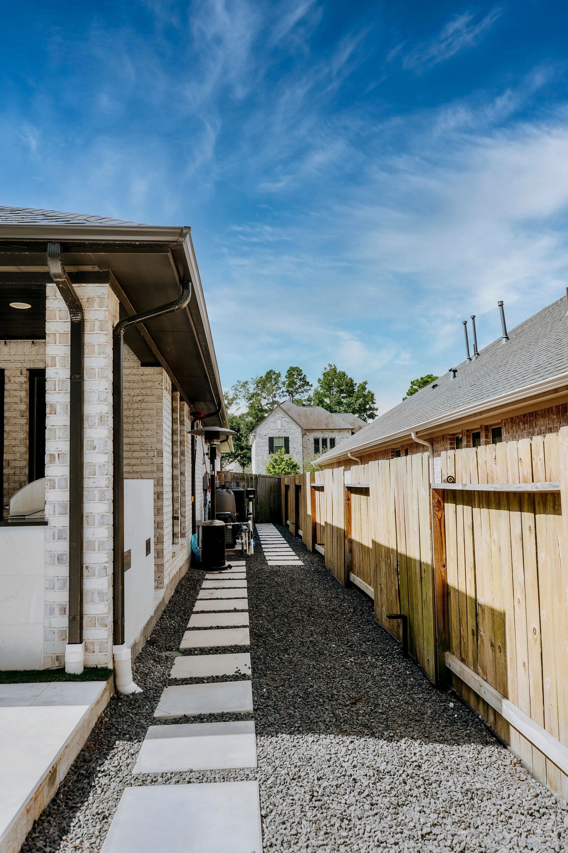 Narrow outdoor walkway between brick buildings and a wooden fence, with stepping stones over gravel under a blue sky