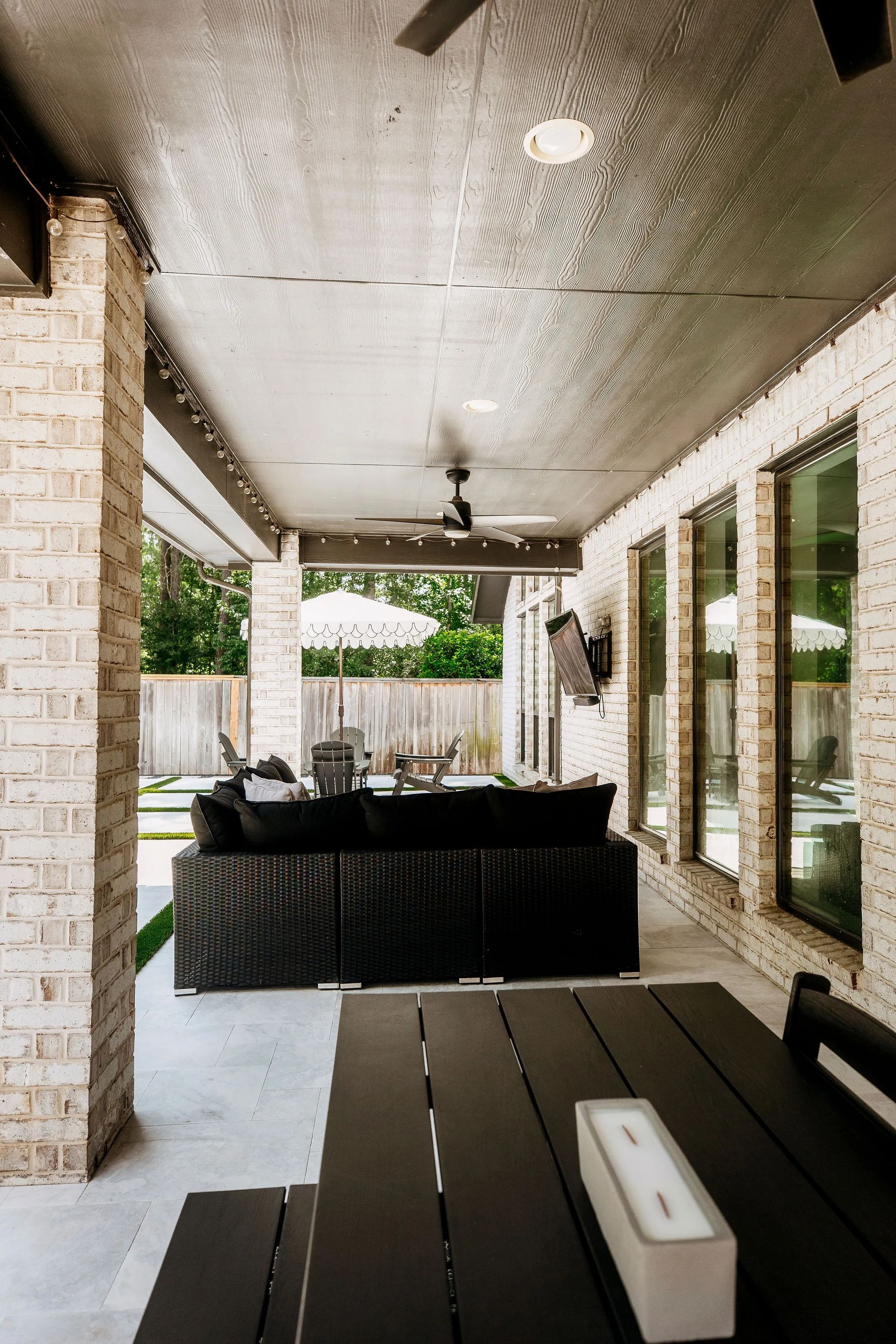 Covered patio with black outdoor sofa, dining table, and brick columns beside large windows