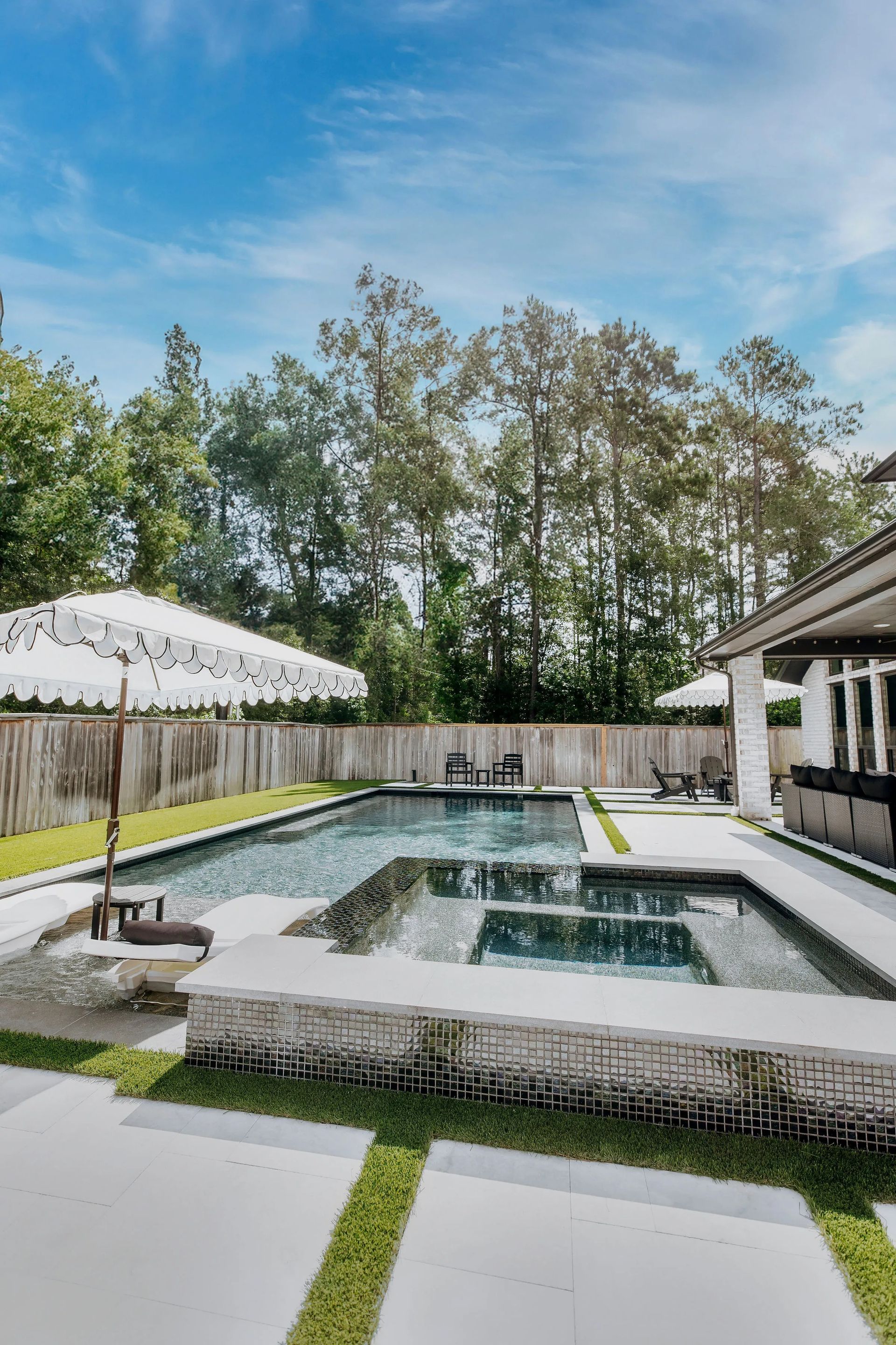 Modern backyard with a raised pool, white patio, lounge chairs, and trees under a blue sky