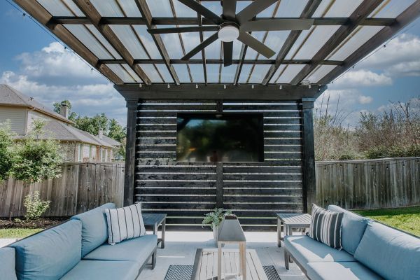 Covered patio with sectional seating, coffee table, and outdoor TV in a fenced backyard