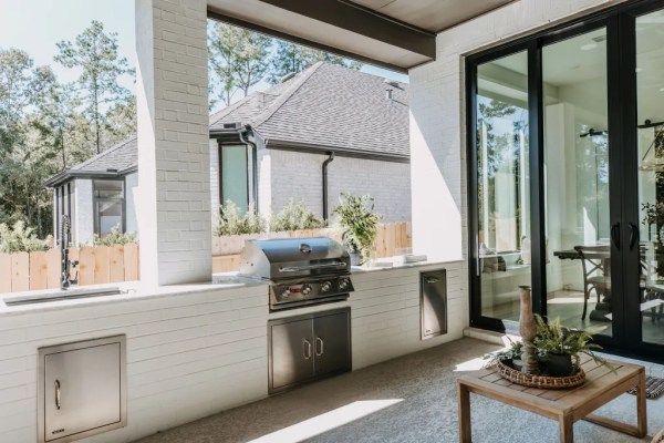Covered patio with built-in grill, white columns, and view of a neighboring house through open windows