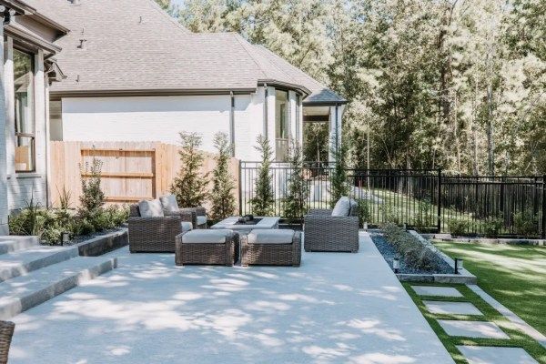 Modern backyard patio with gray outdoor seating, stone steps, and a fenced lawn beside a house