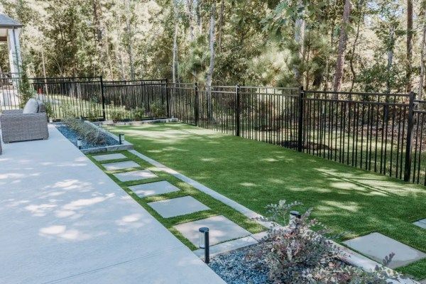 Backyard patio with stepping stones across a green lawn, black fence, and tall trees beyond