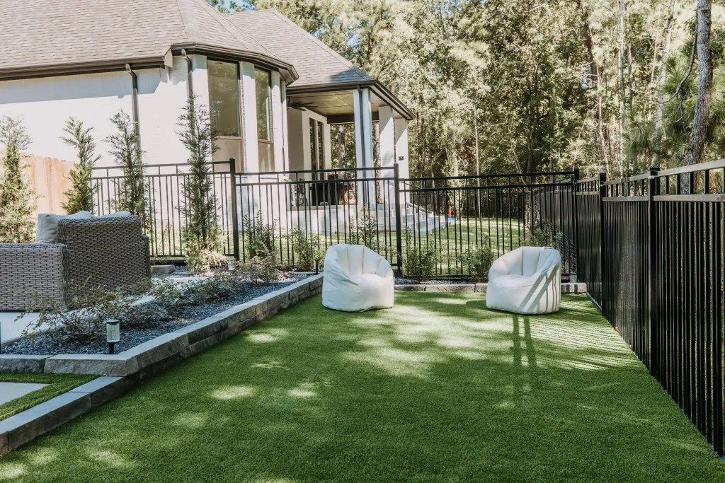 Backyard patio with green lawn, two white chairs, and a black fence beside a house and trees