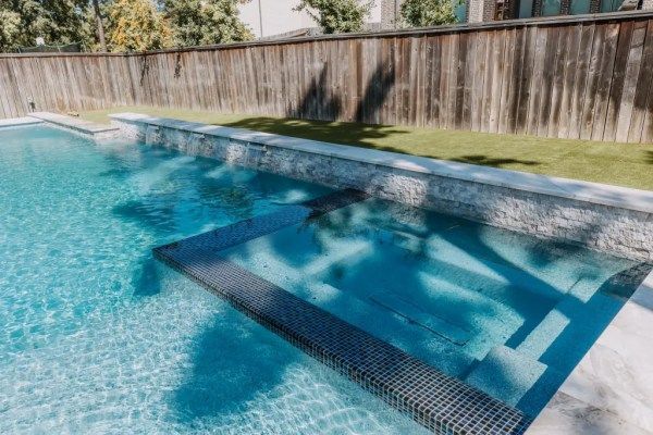 Backyard swimming pool with a shallow ledge, blue water, and wooden fence backdrop