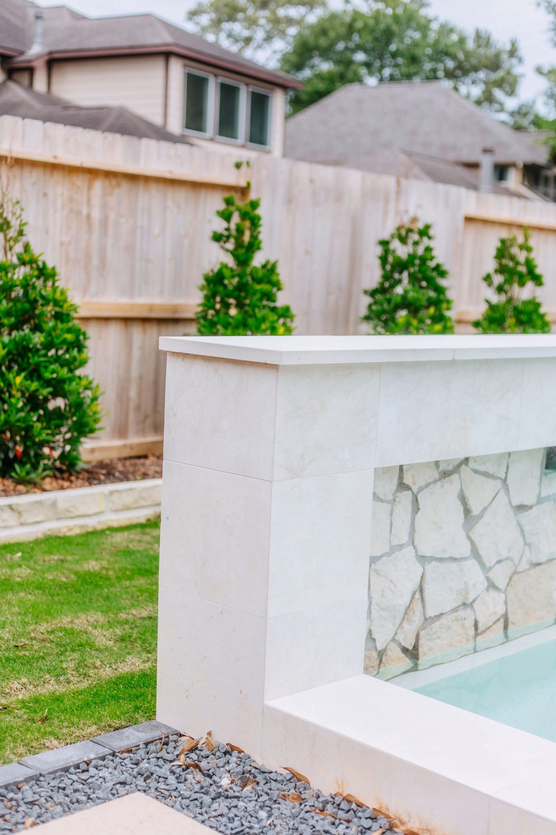 Backyard pool with stone-trimmed concrete edge, green lawn, shrubs, and wooden fence