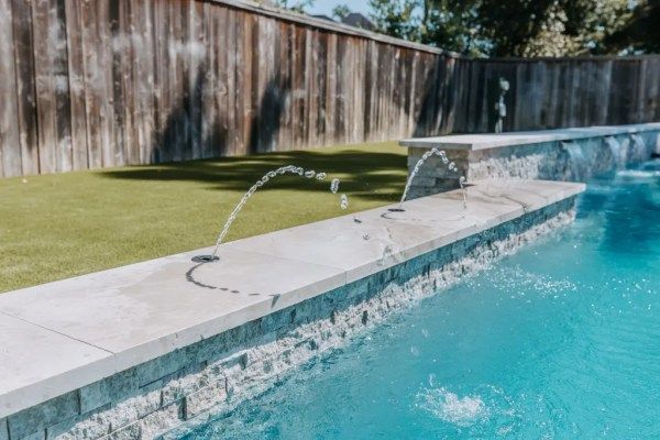 Swimming pool edge with two water fountains spraying into turquoise water beside a grassy lawn and wooden fence