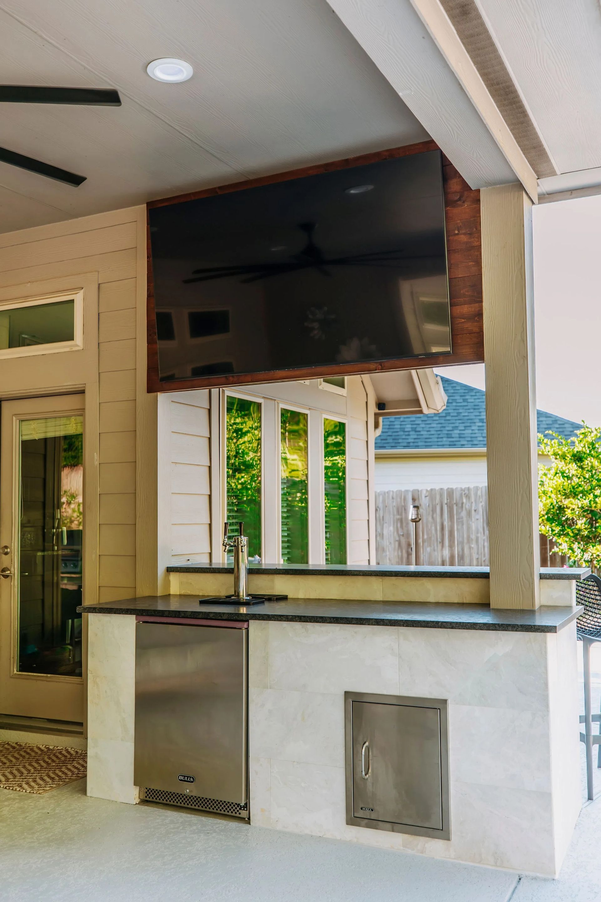 Outdoor TV mounted above a backyard bar with a black countertop and white cabinets under a covered patio