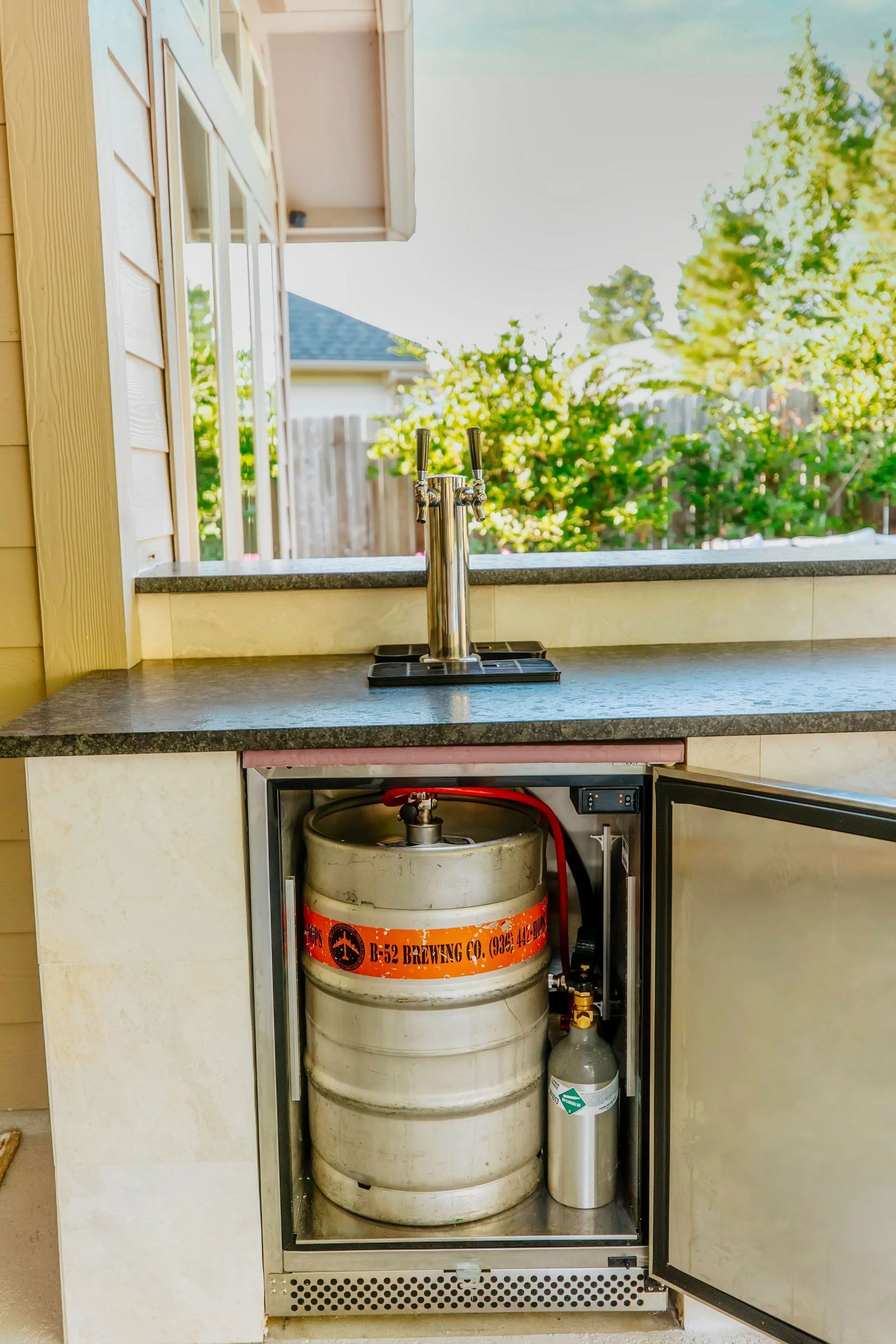 Open under-sink cabinet with gas cylinder and small bottle beneath a countertop outdoors