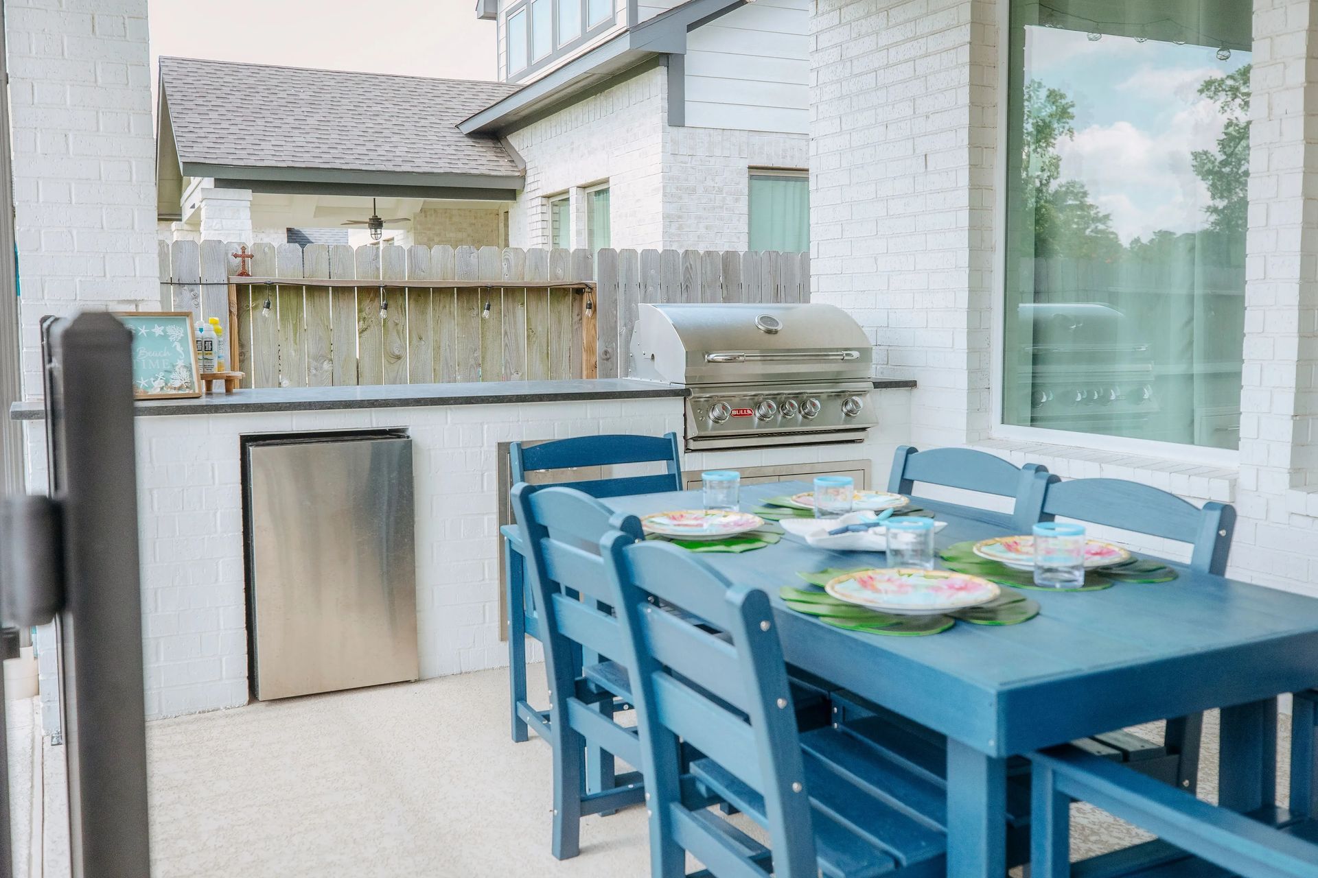 Blue patio dining set beside a grill on a small outdoor deck with a fenced yard beyond