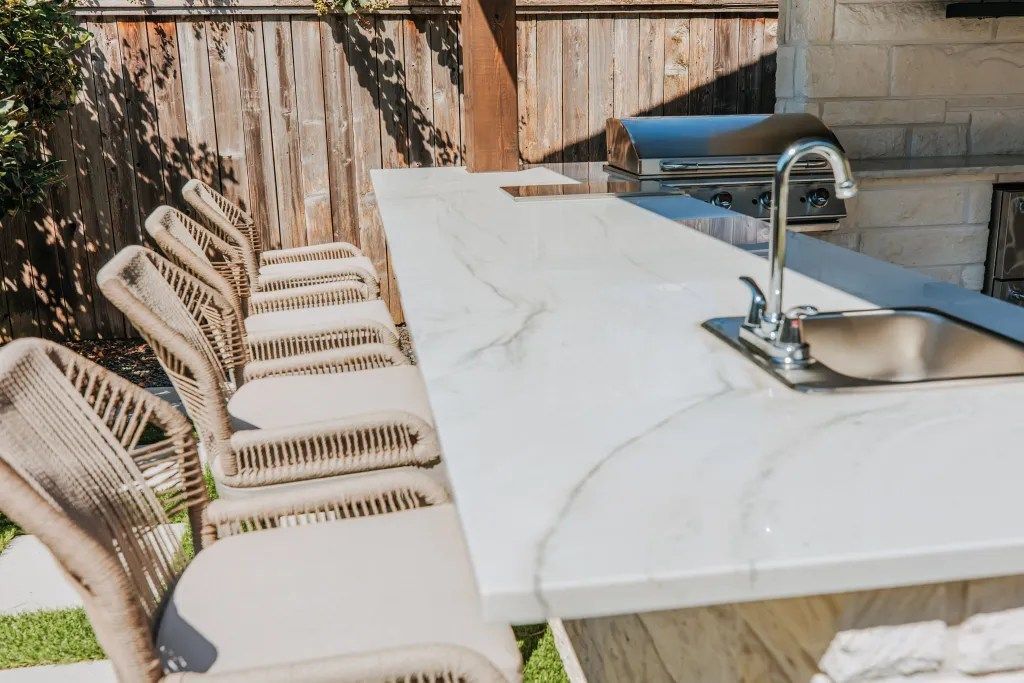 Outdoor patio with white dining table, woven chairs, and a metal sink beneath a wooden fence.