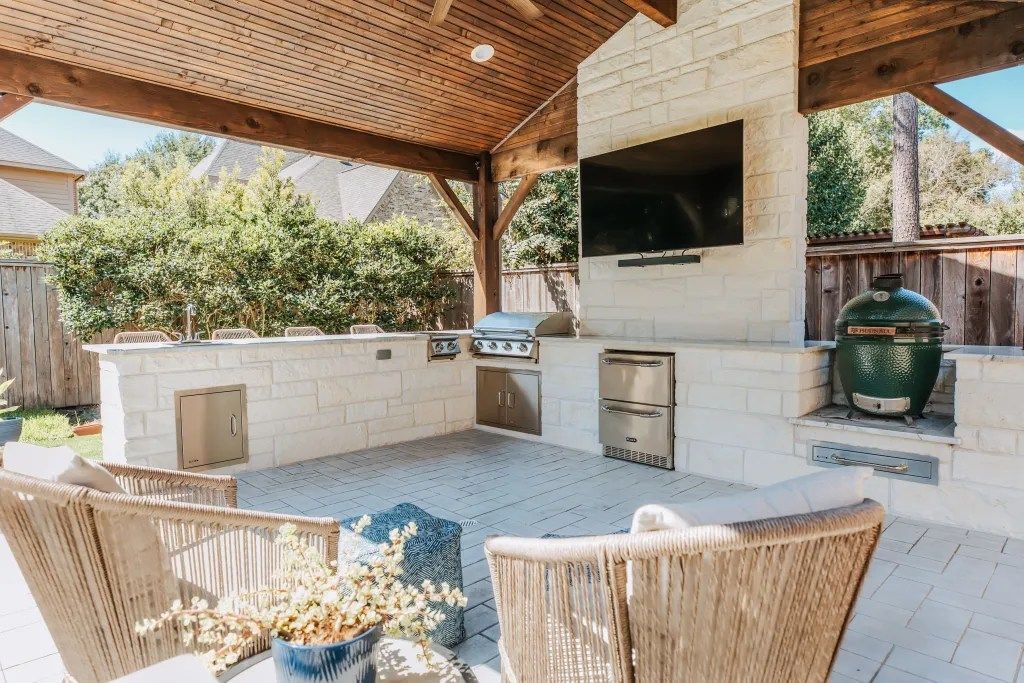 Covered outdoor patio with built-in grill, white stone counter, wicker chairs, and wall-mounted TV