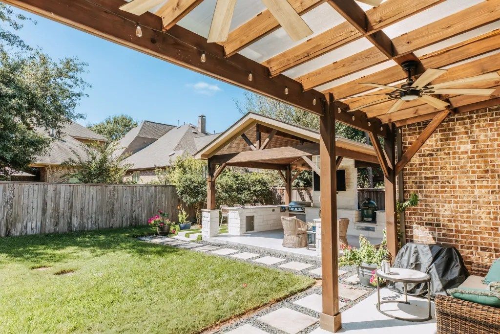 Covered backyard patio with wooden beams, lawn, stone path, outdoor seating, and a grill by a brick wall.