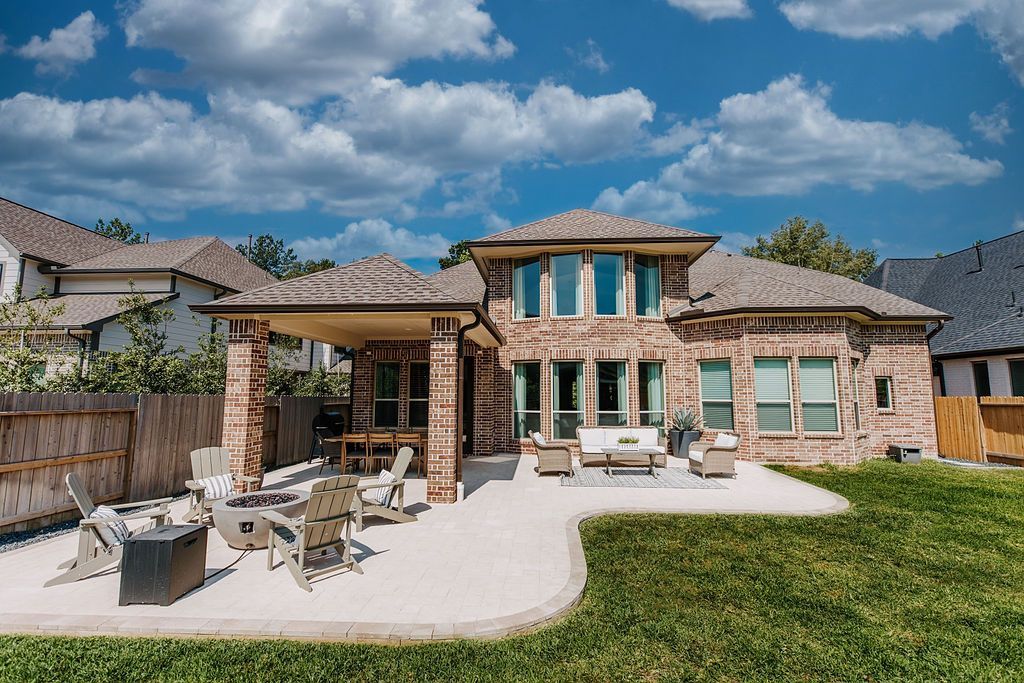 Modern two-story stone house with patio furniture, green lawn, and blue sky with clouds