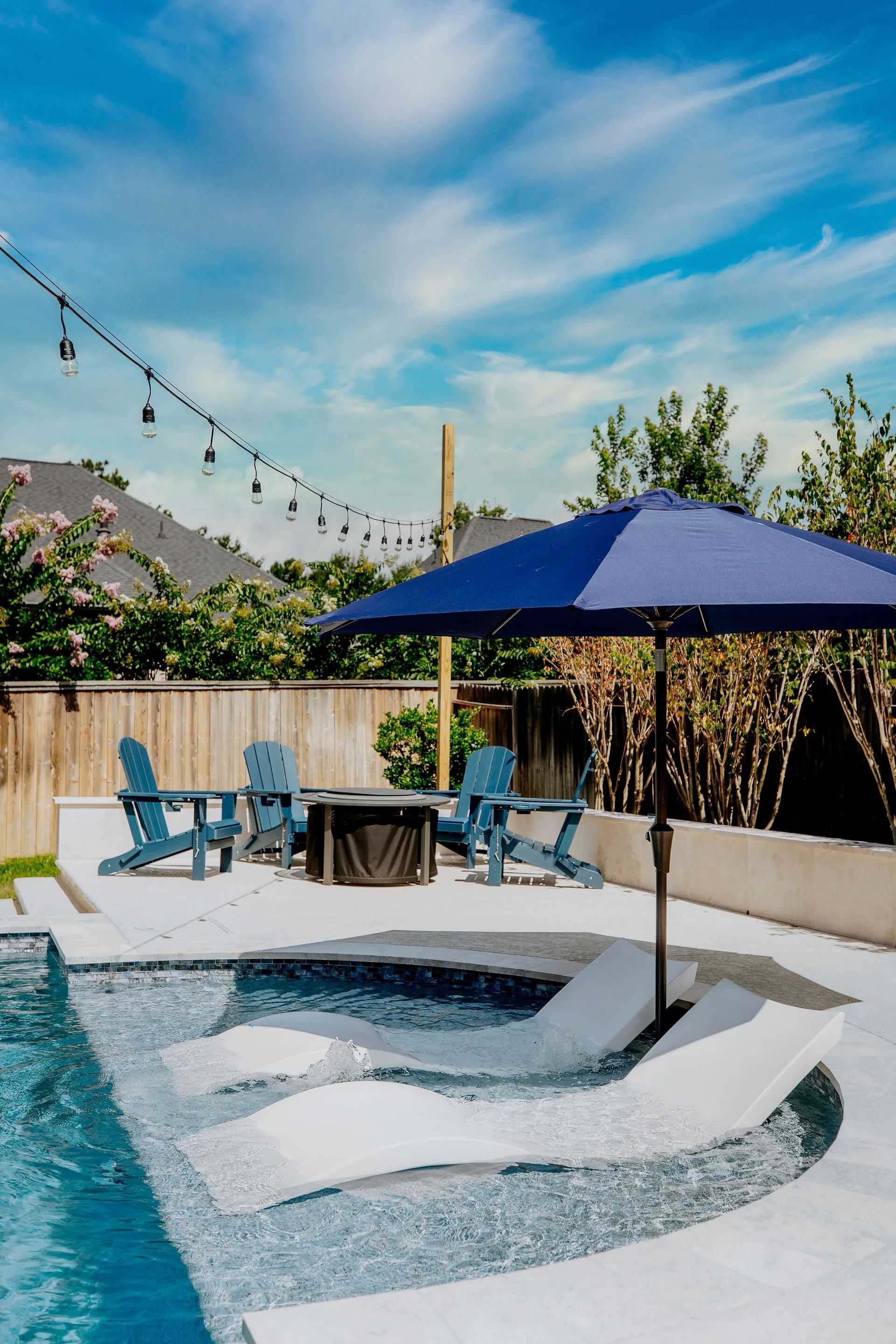 Backyard pool with white lounge chairs, blue umbrella, string lights, and mountain view under a sunny sky