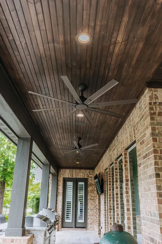 Covered porch with wood-paneled ceiling, ceiling fan, brick wall, and glass double doors.