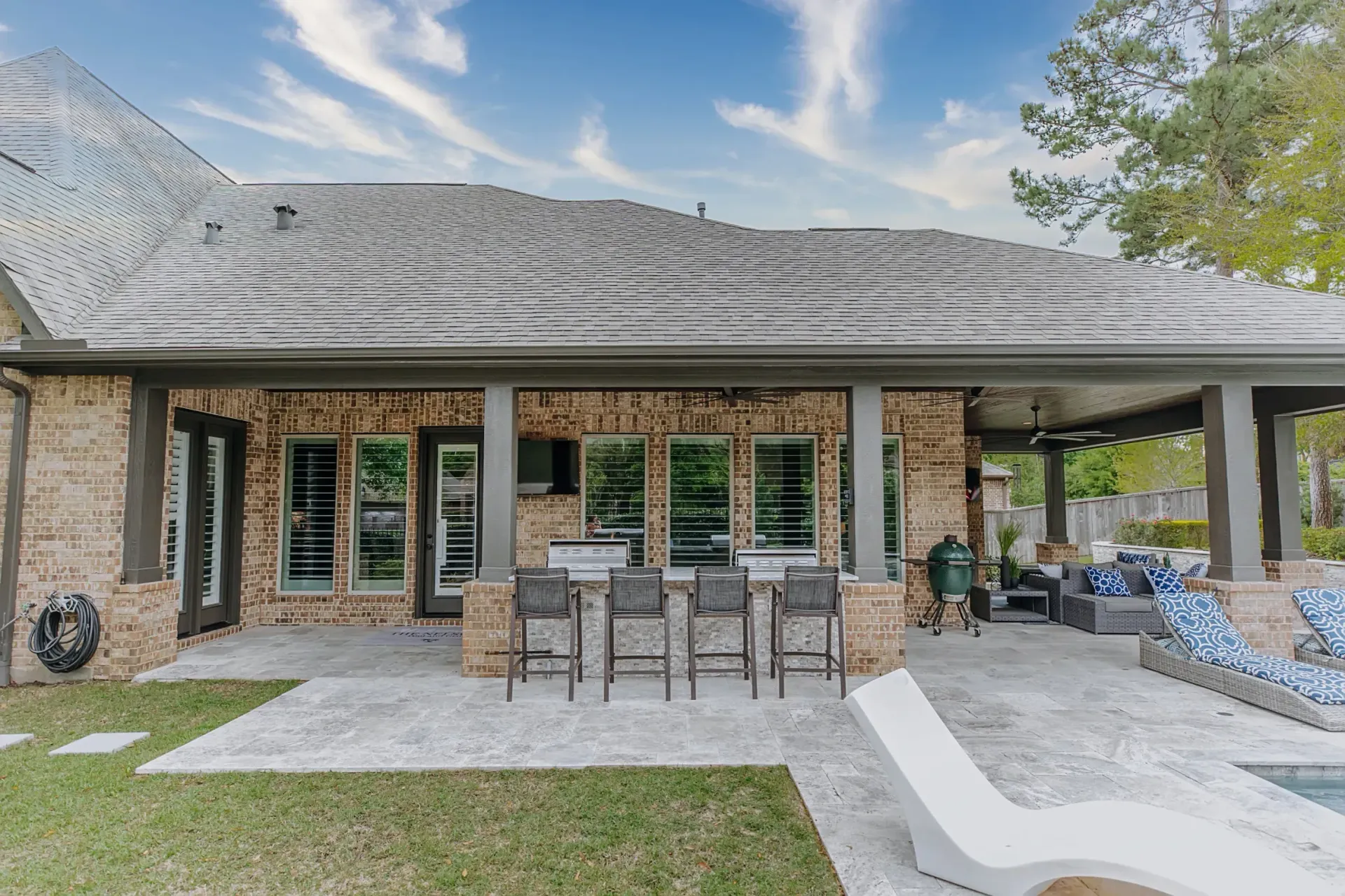 Covered stone patio with outdoor dining table, chairs, and lounge seating beside a house and pool.