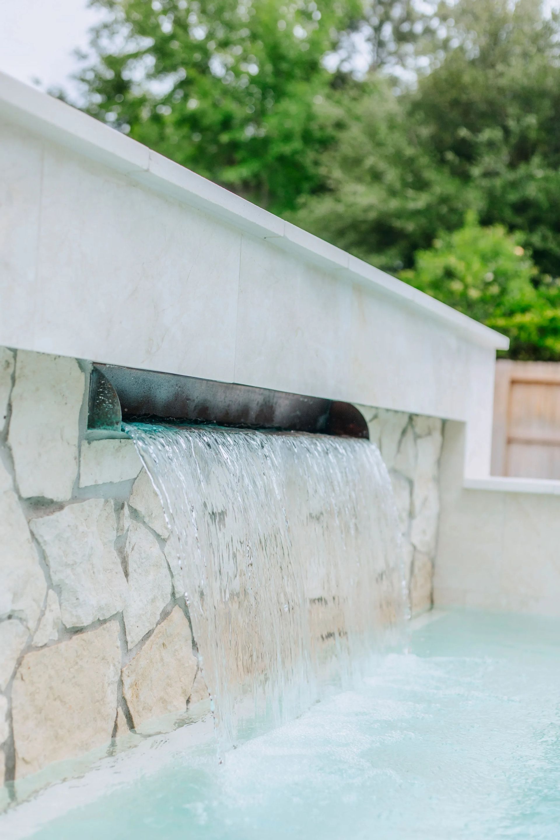 Modern stone water wall with a narrow spout spilling into a light blue pool outdoors