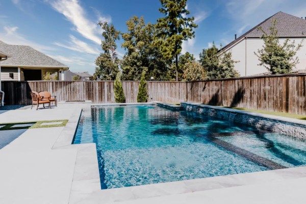 Backyard swimming pool with blue water, white deck, wooden fence, and lounge chair under a sunny sky